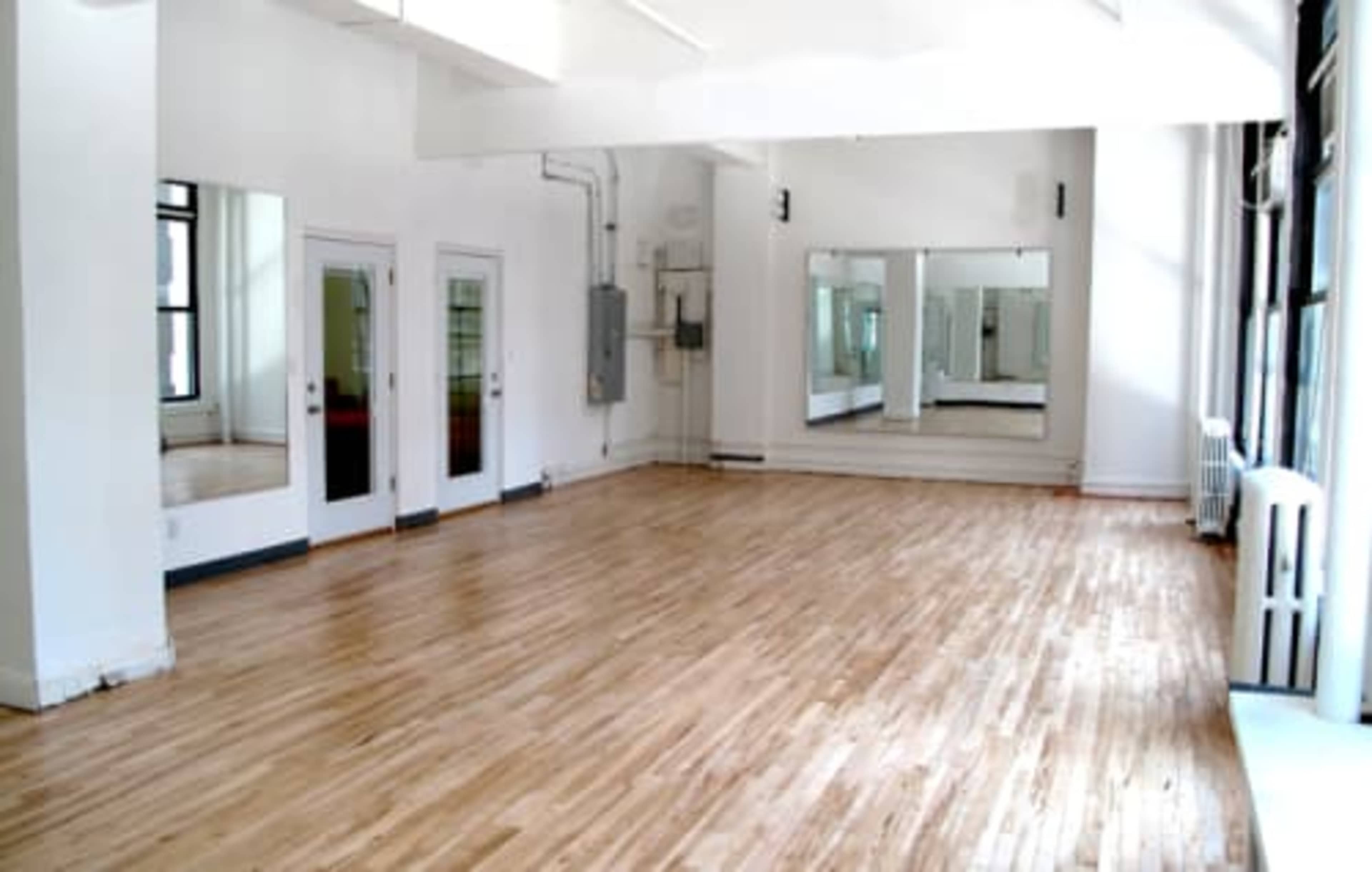The image depicts a spacious, empty room with hardwood floors and large mirrors on the walls.