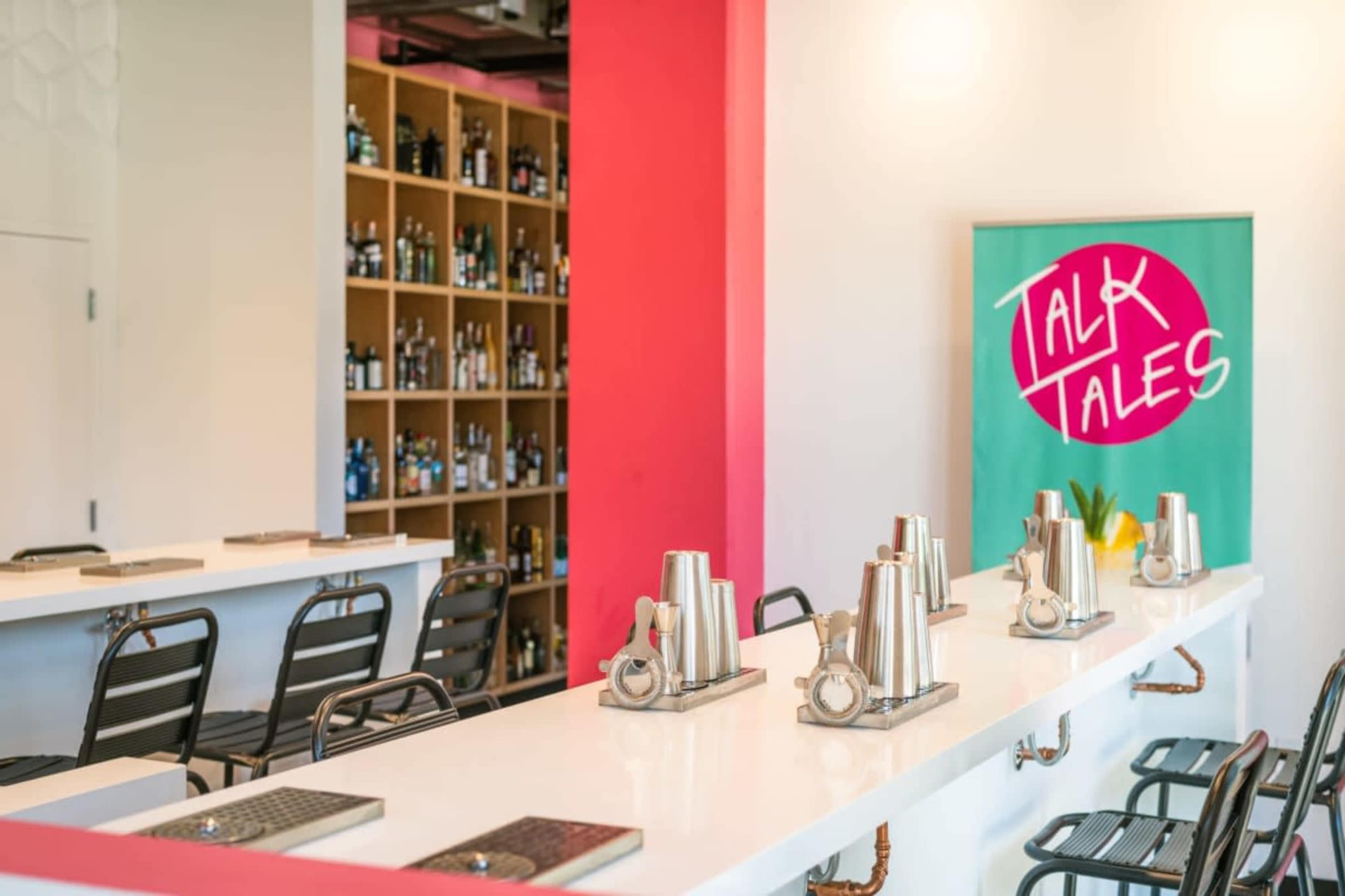 The image shows a modern bar with a white countertop, stainless steel cocktail shakers arranged on it, black chairs lined up in front, and a wall featuring colorful shelves stocked with bottles and a vibrant sign that reads "TALK TALES."