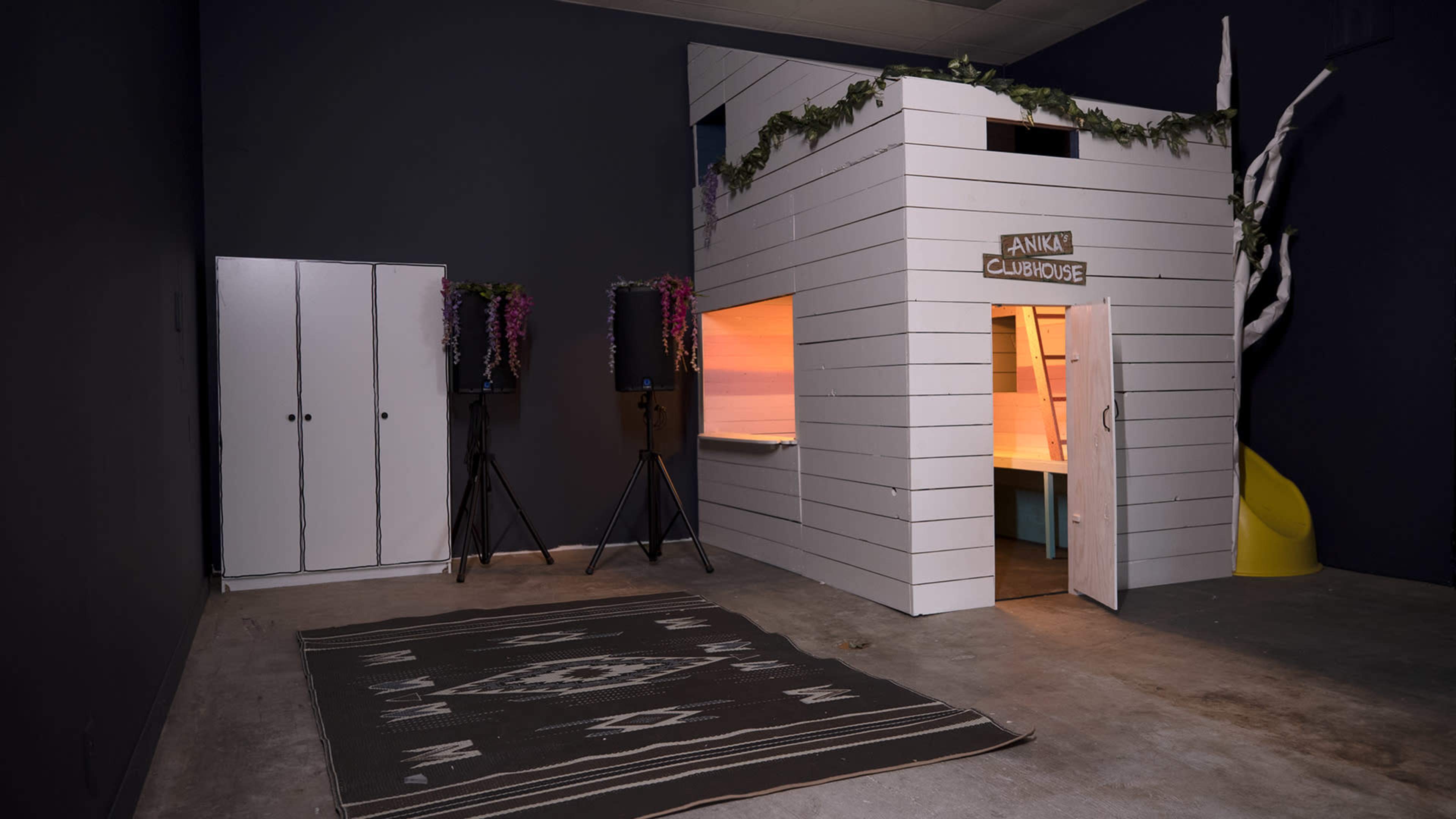 A small, modern clubhouse structure made of white wood, accompanied by speakers, a wardrobe, and a patterned rug on the floor.