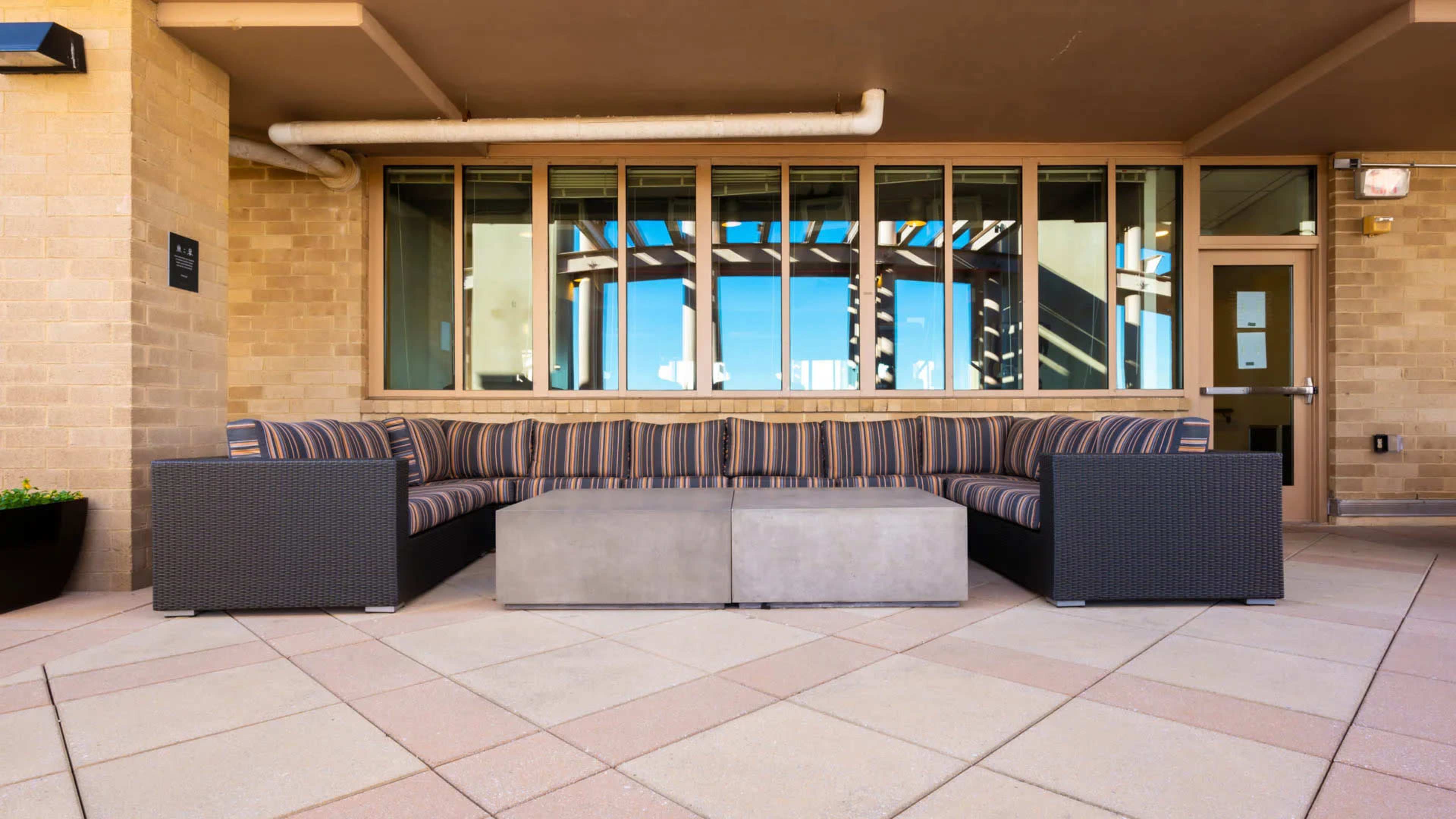A sectional outdoor sofa with striped upholstery and a concrete coffee table is situated on a tiled patio with large windows nearby.