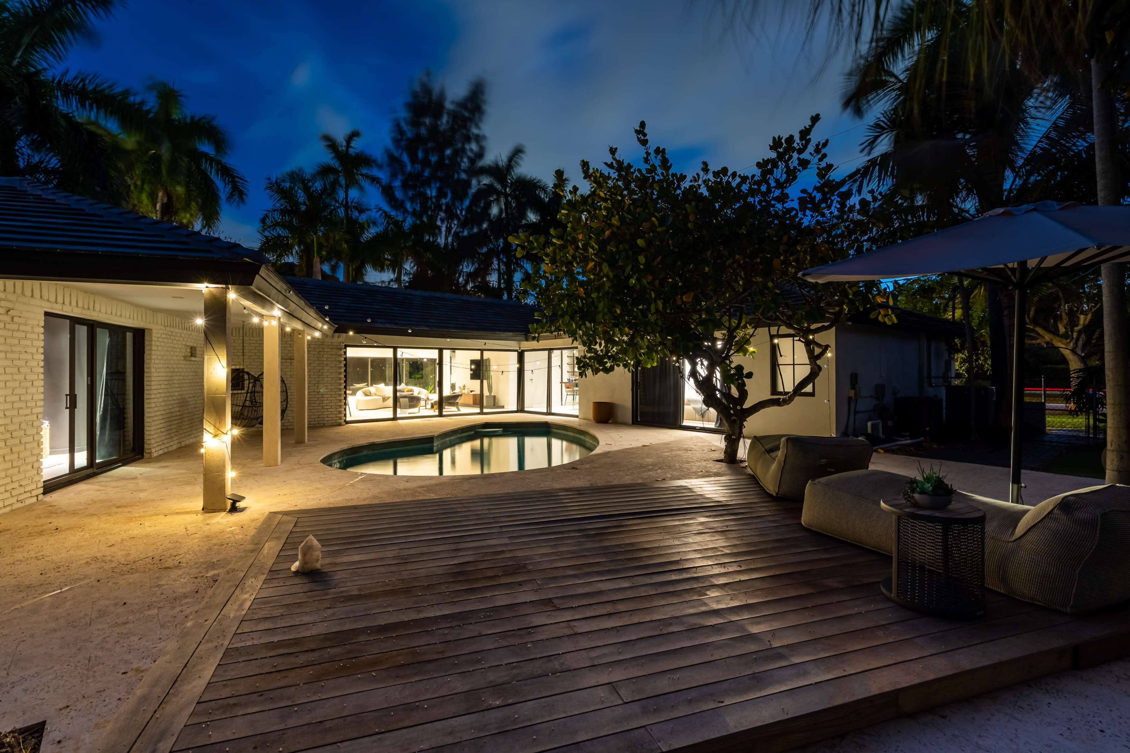 The image shows a modern outdoor living space with a circular pool surrounded by a wooden deck, illuminated by soft lighting against a backdrop of palm trees at night.