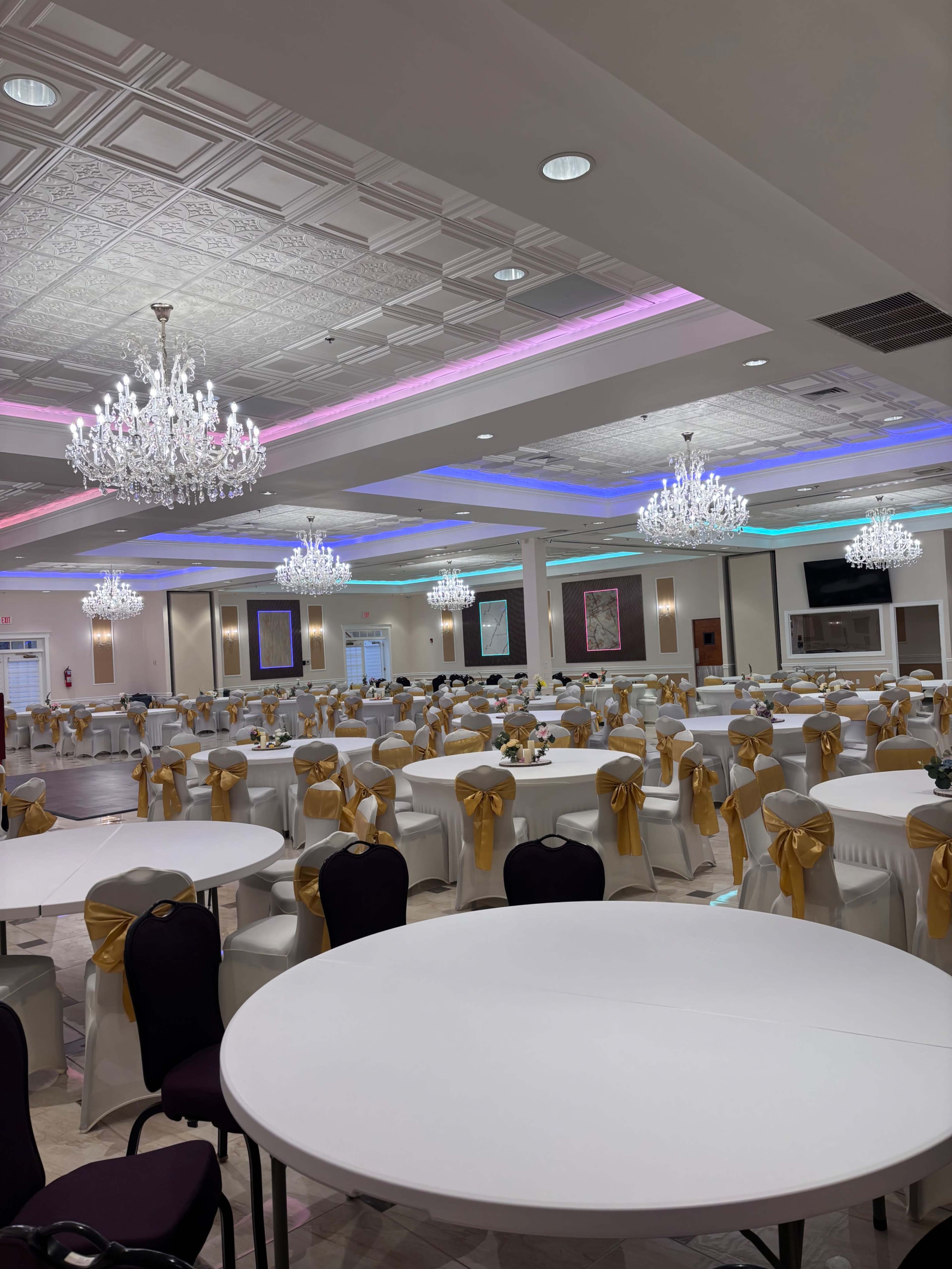 A spacious banquet hall features numerous round tables with white tablecloths and yellow sashes, illuminated by multiple chandeliers and colorful ceiling lights.