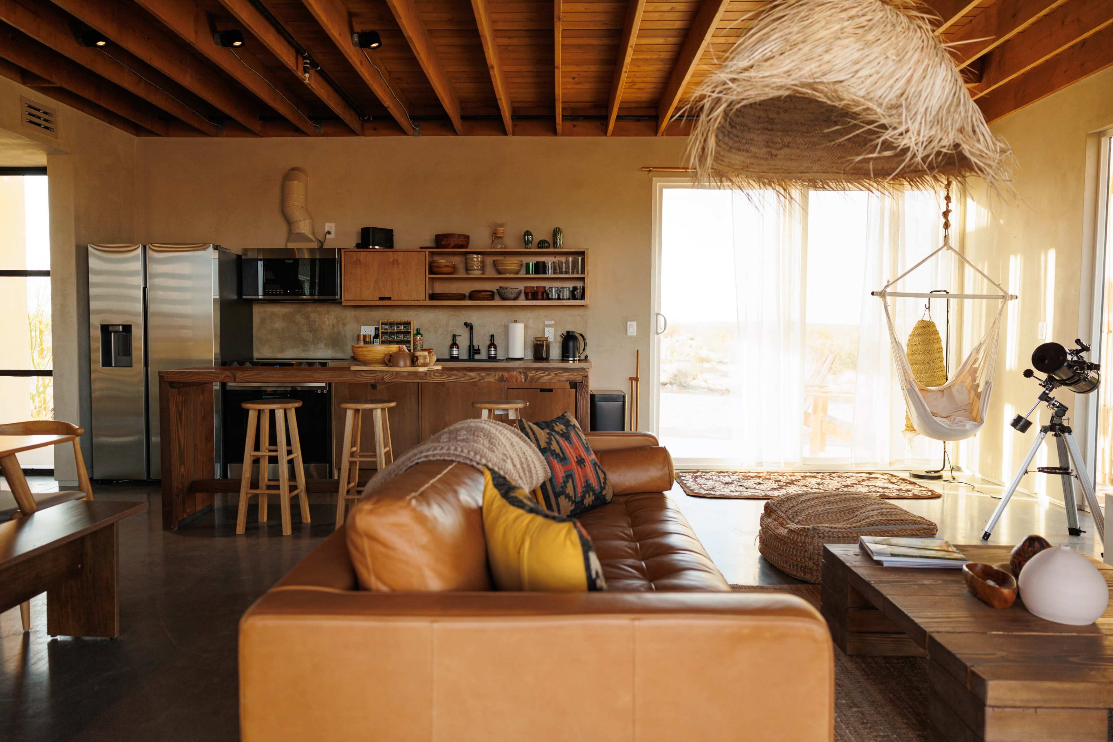 The image shows a modern, open-concept living space with a kitchen area, seating on a leather sofa, and a hanging chair near large windows.