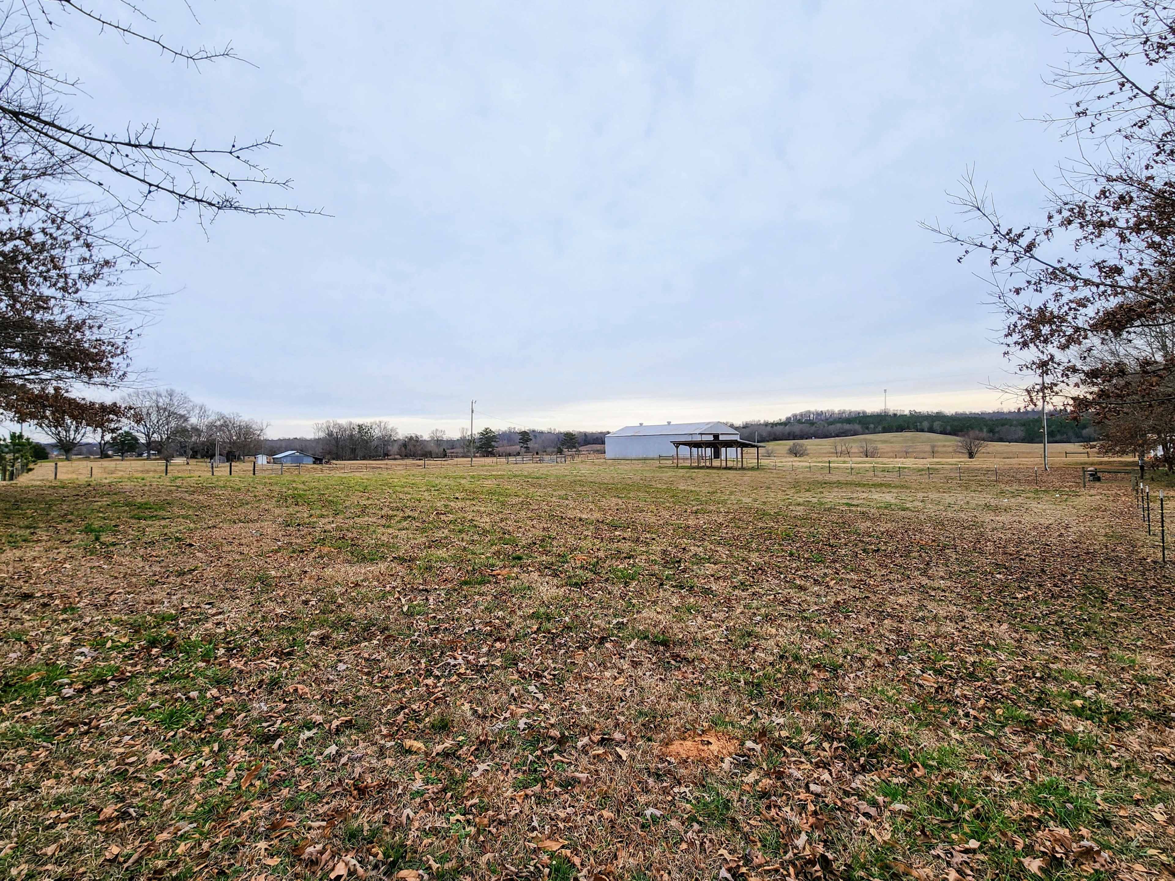 A wide open field with sparse trees and a few structures in the distance under a cloudy sky.