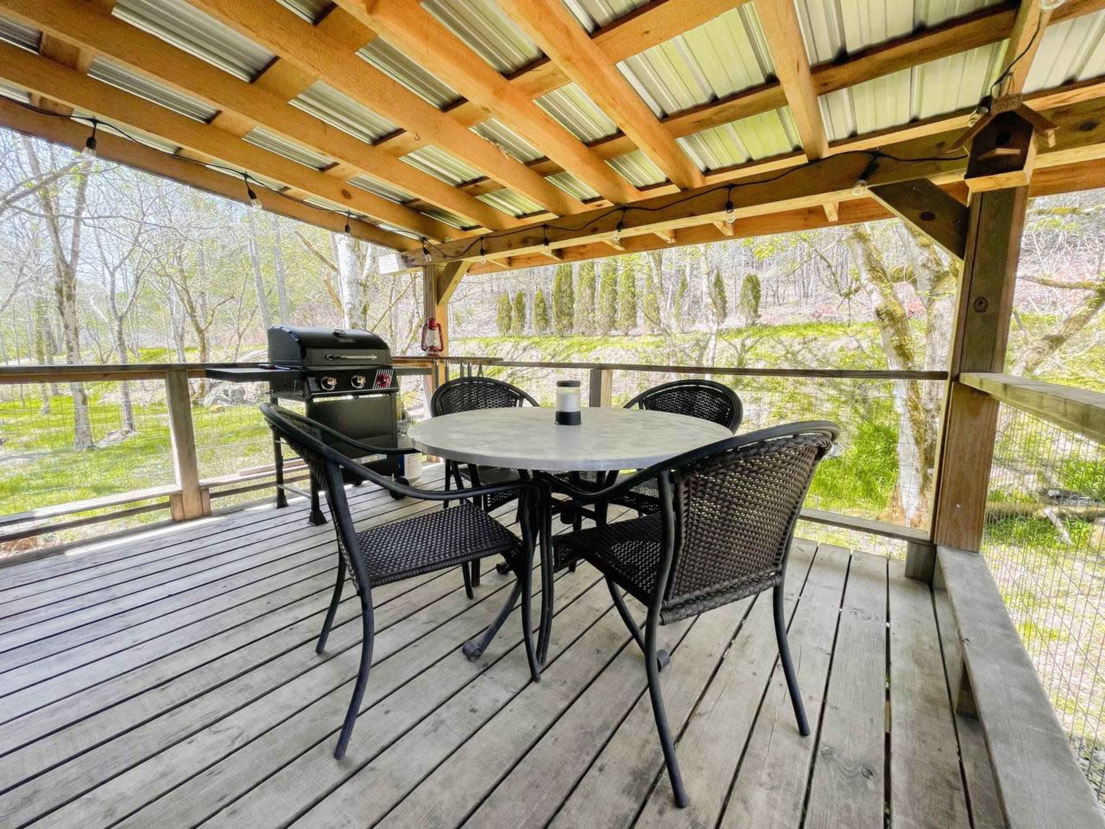 The image shows a wooden deck with a round table and four black chairs, alongside a grill and surrounded by trees.