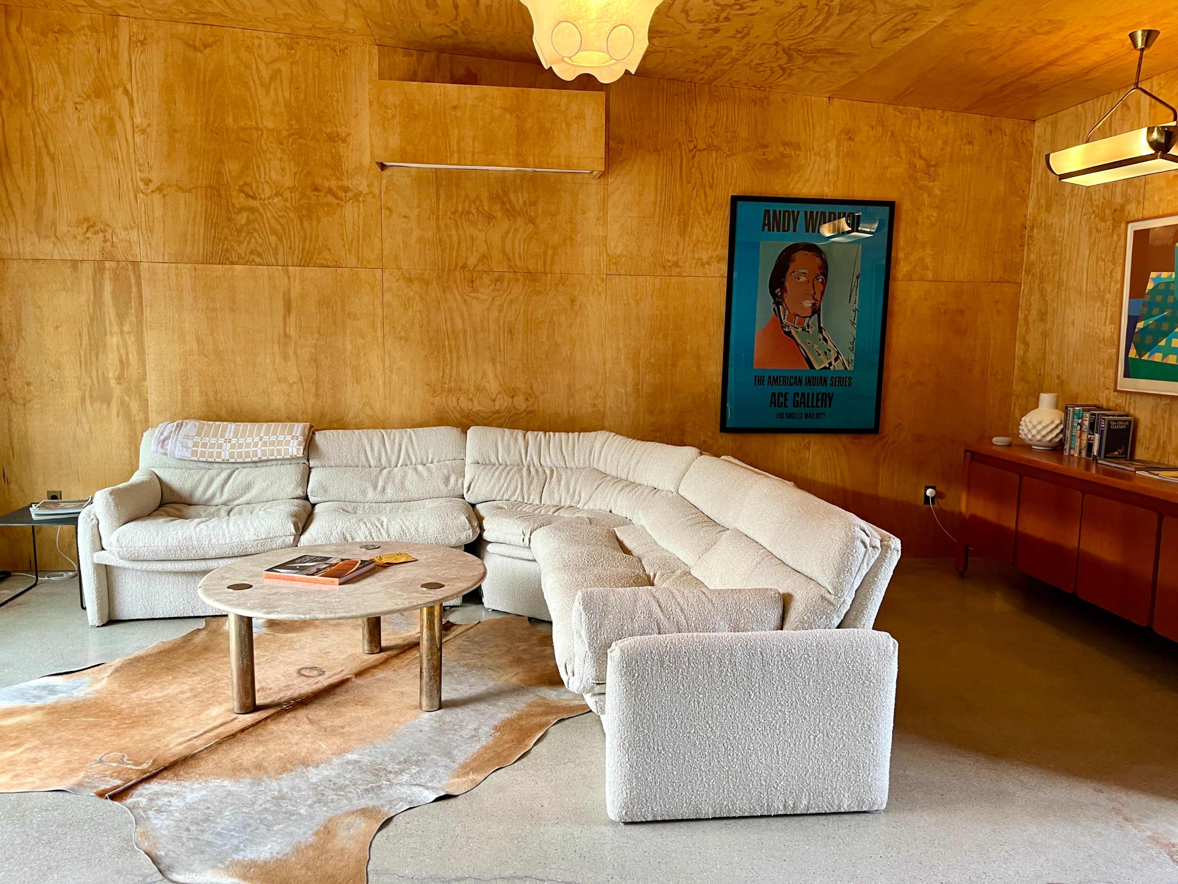 A cozy living room with a large, curved sectional sofa, a round coffee table on a cowhide rug, and a wooden sideboard beneath a colorful poster.
