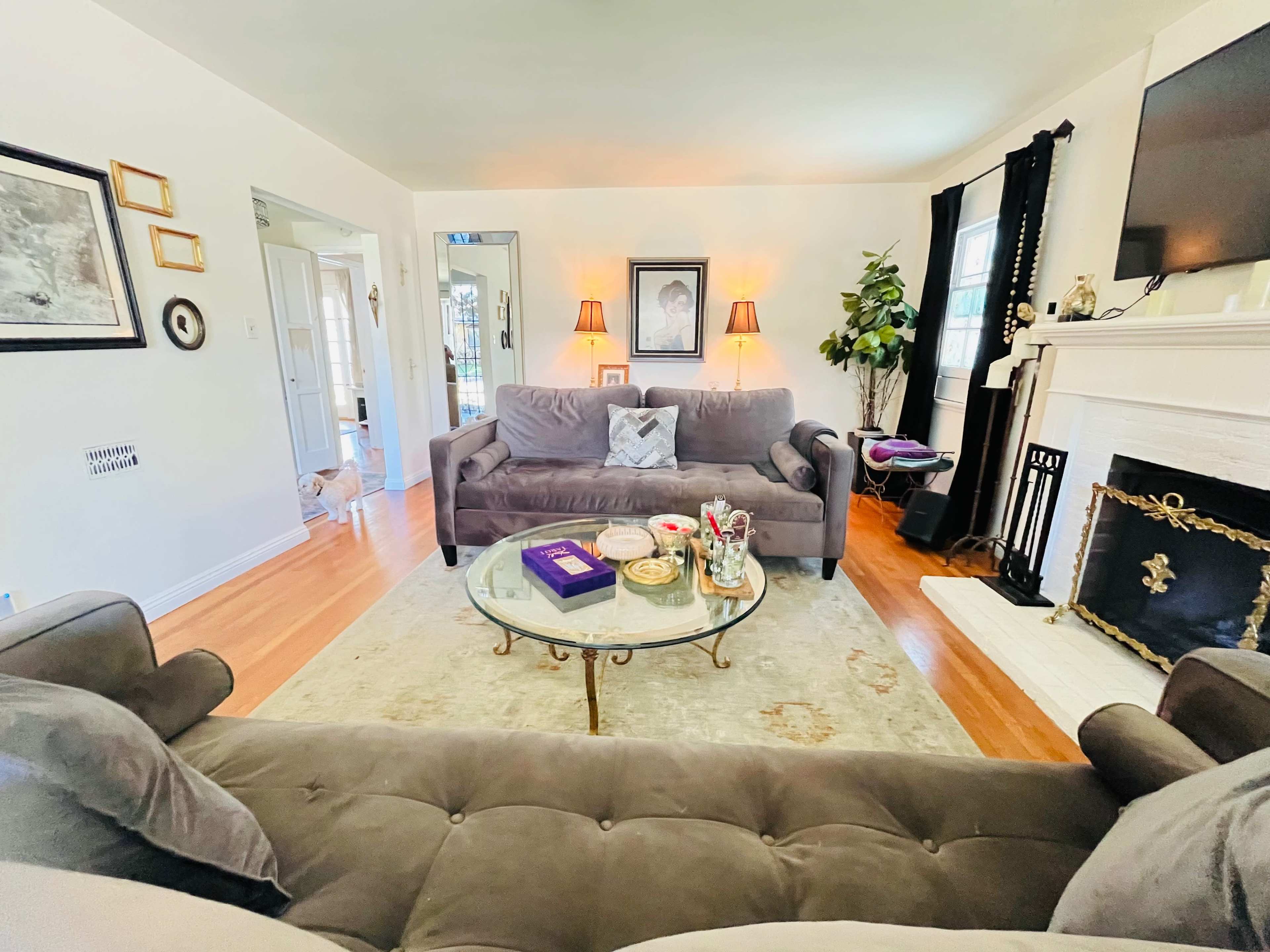 The image shows a living room with two gray sofas facing a glass coffee table on a light-colored rug, and a fireplace adorned with decorative items.