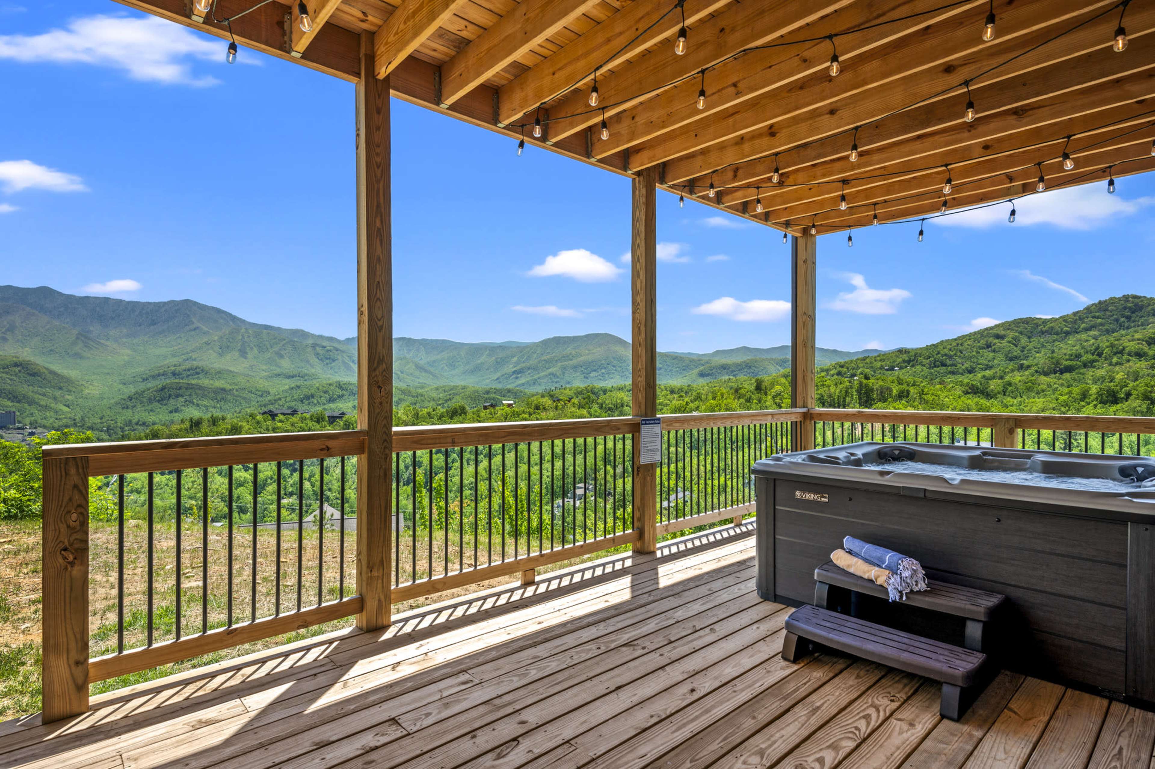 A wooden patio with a hot tub, overlooking green mountains under a clear blue sky.