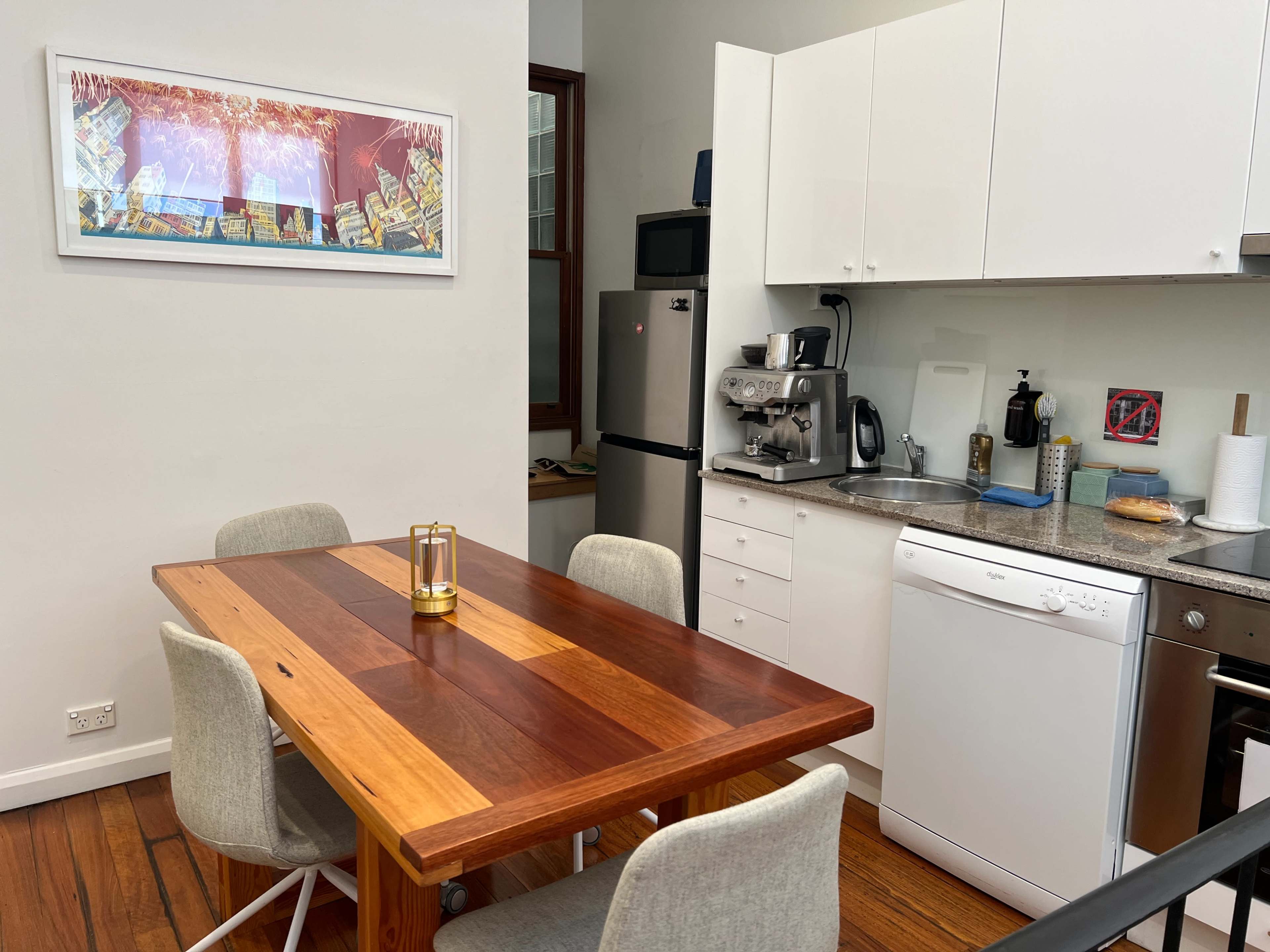A modern kitchen with a wooden dining table, chairs, a sink, and various kitchen appliances against a light-colored wall.