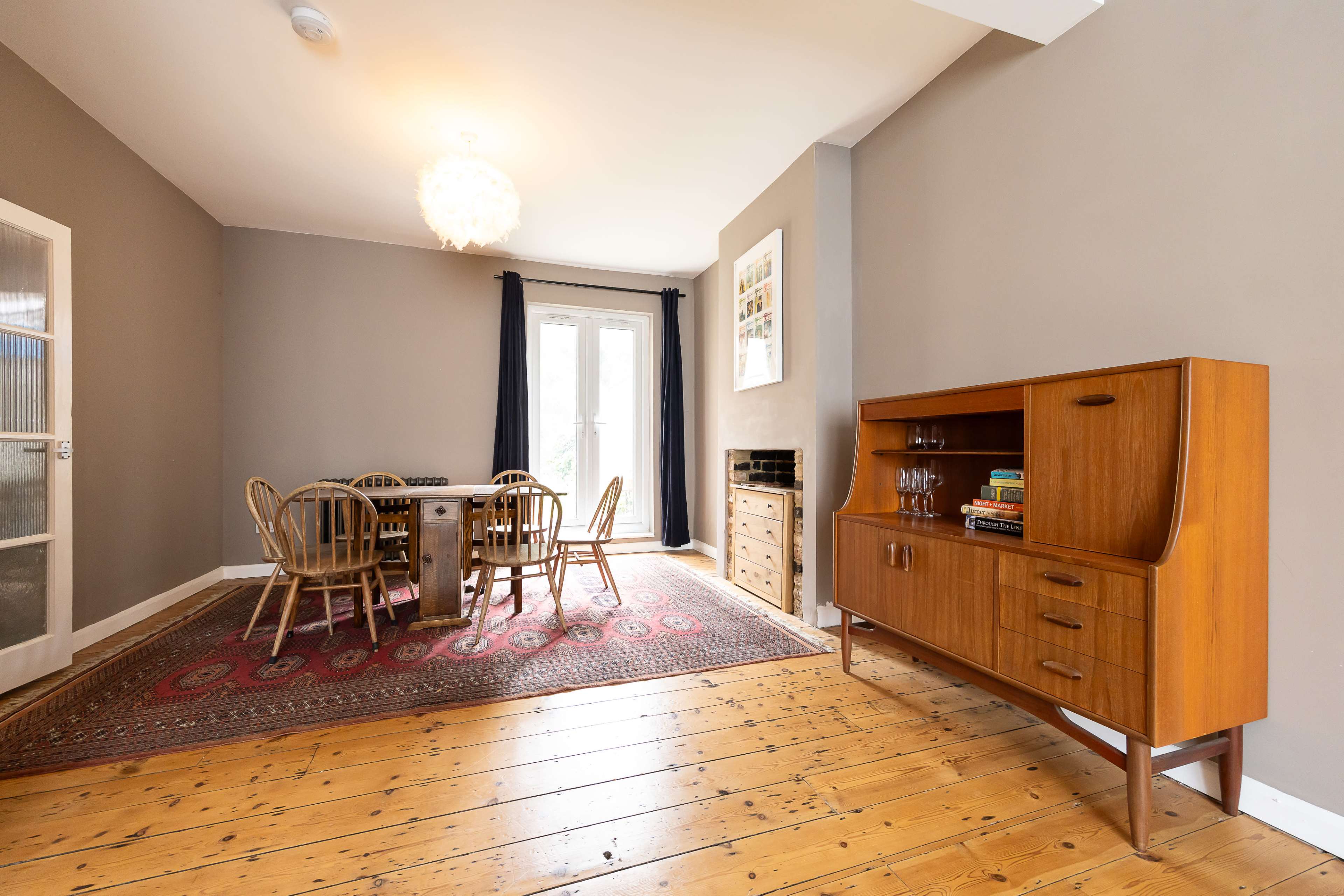 The room features a dining area with a wooden table and chairs, a sideboard, and a decorative rug on wooden flooring, alongside a large window.