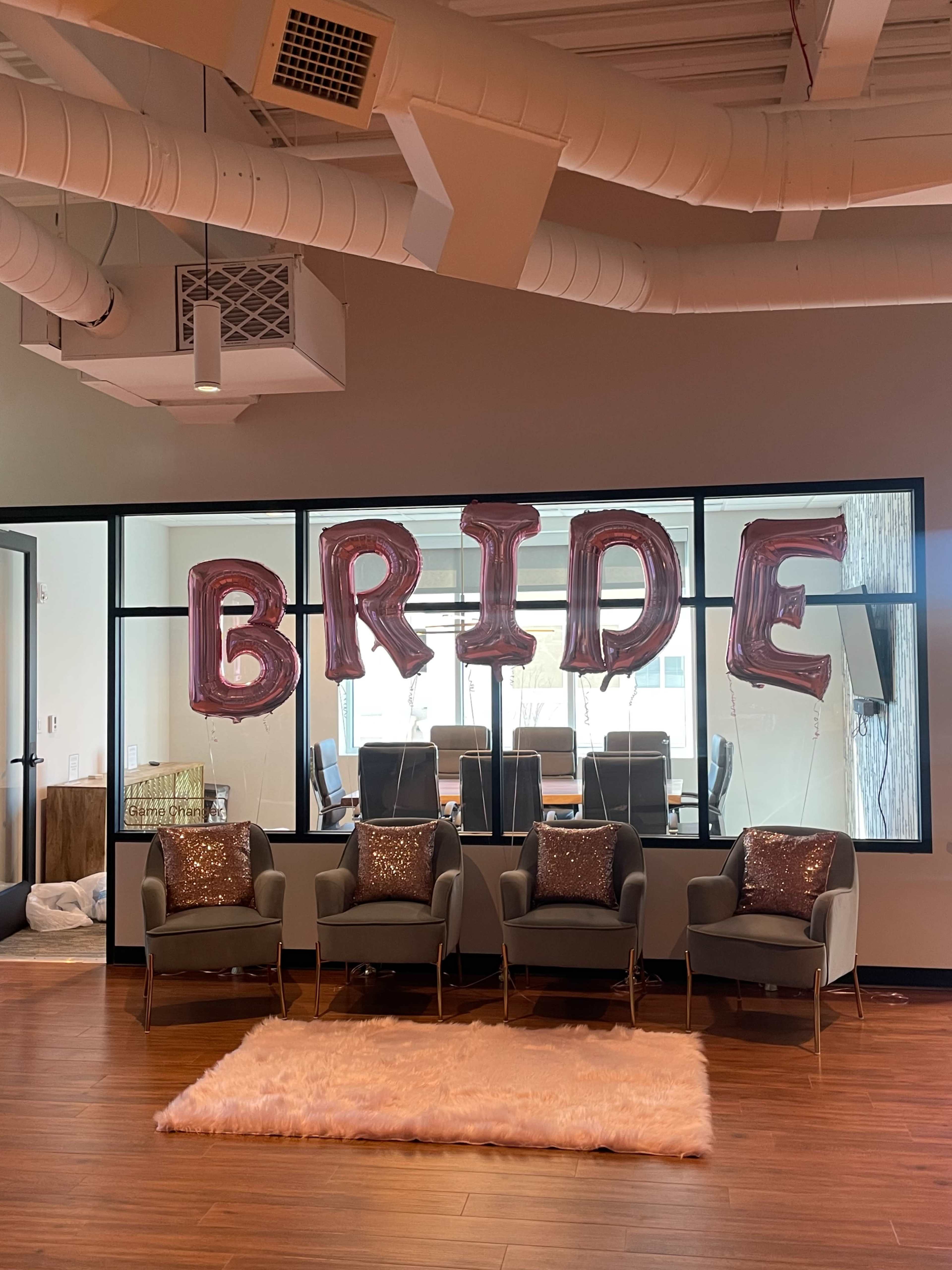 A set of four chairs with sequined cushions is positioned in front of large balloon letters spelling "BRIDE" against a backdrop of a glass conference room.