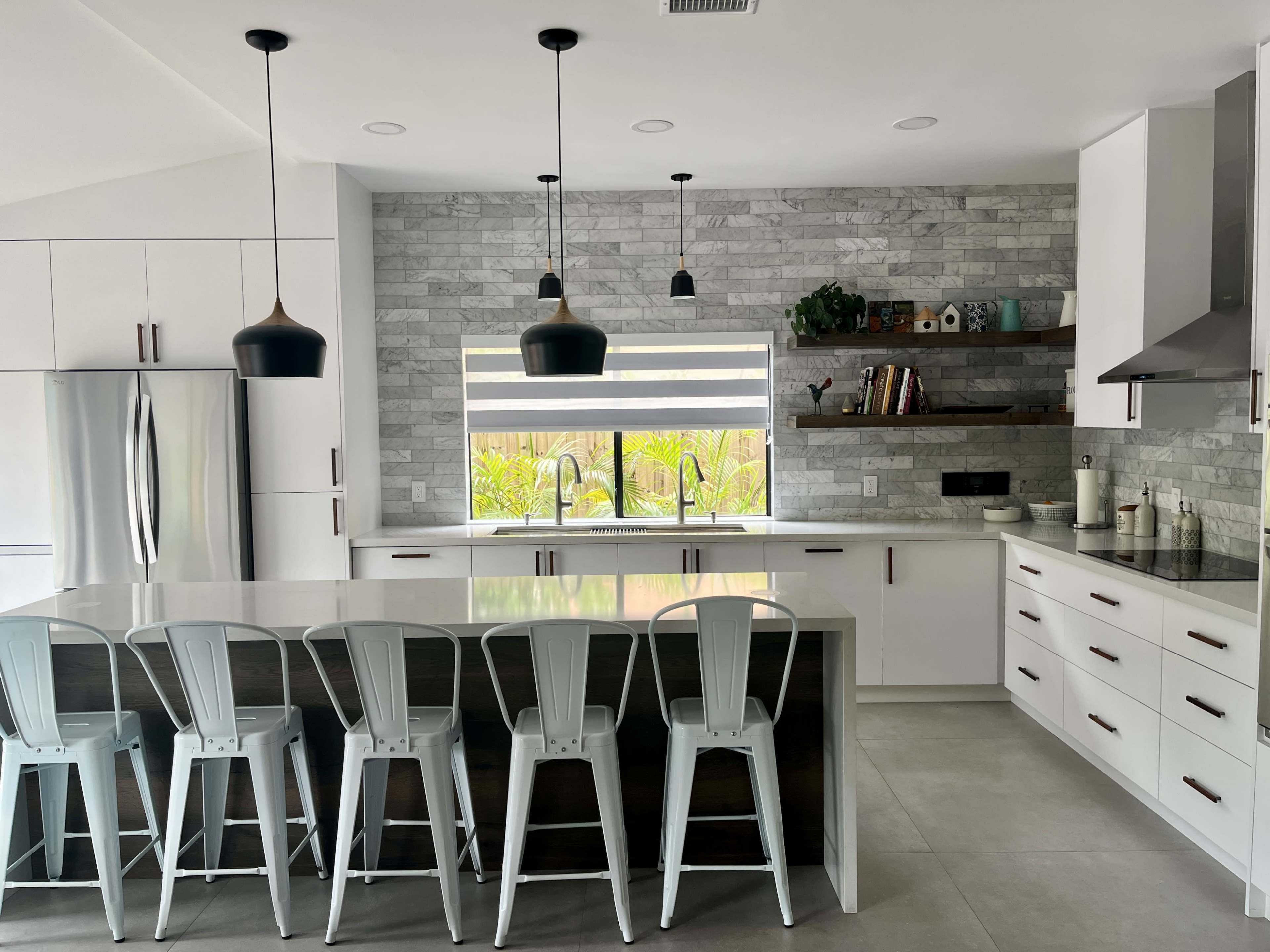A modern kitchen features a large island with metal bar stools, stainless steel appliances, and a stone accent wall with open shelving.