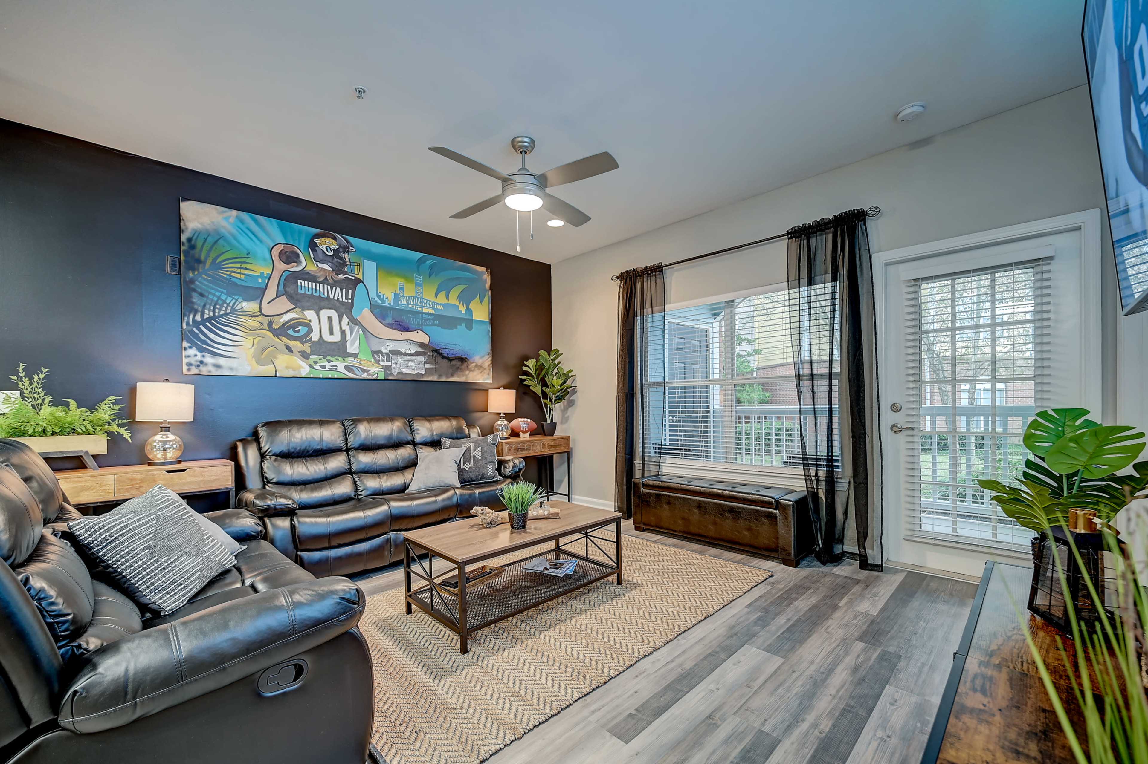 The image shows a living room featuring black leather furniture, a wall mural, potted plants, and a patio door with blinds.