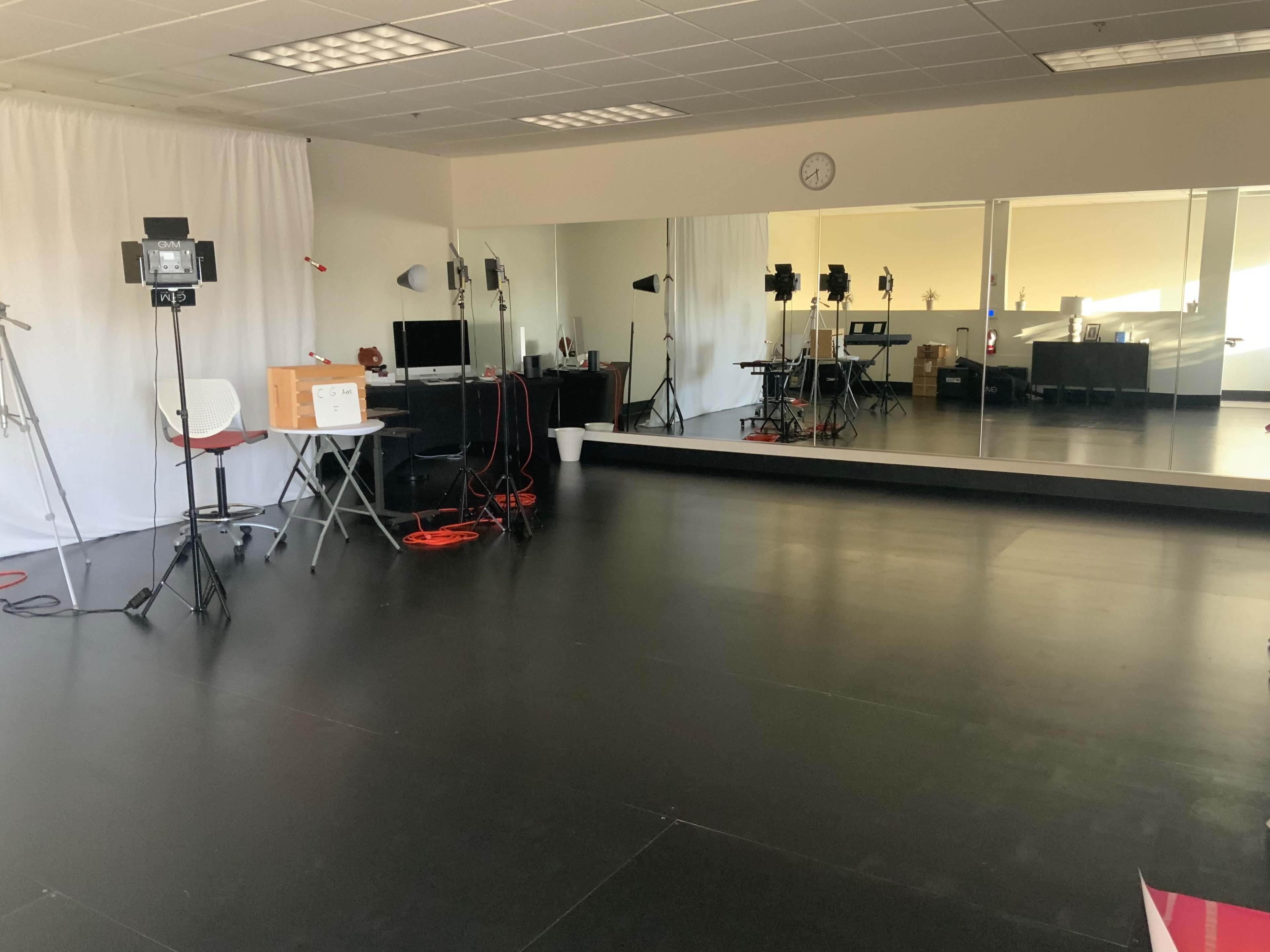 A well-lit studio space featuring a mirrored wall, equipment such as lights and cameras, and minimal furniture including chairs and tables.