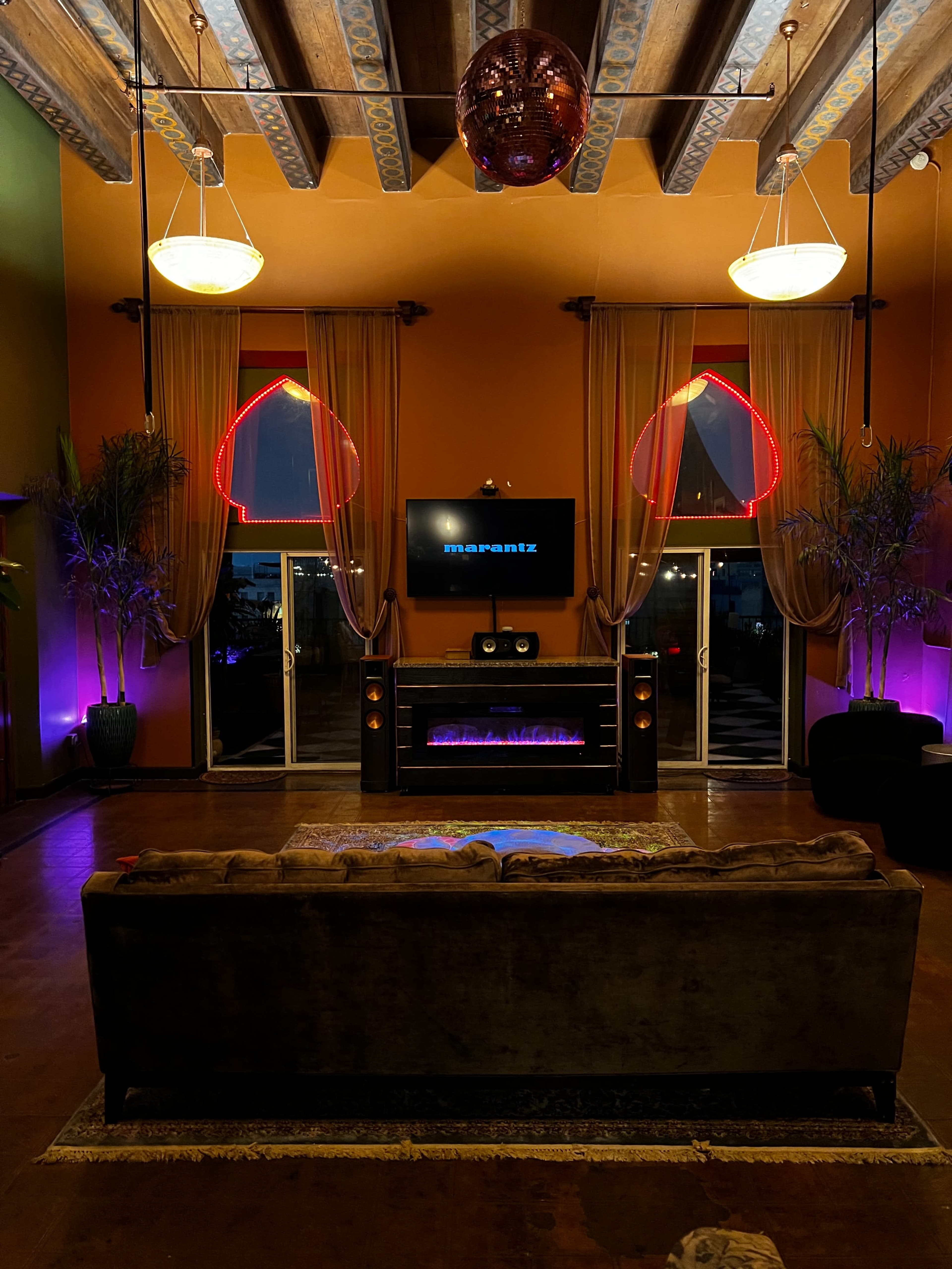A dimly lit lounge area with a velvet sofa facing a wall-mounted television, framed by large windows illuminated with pink and purple lights.