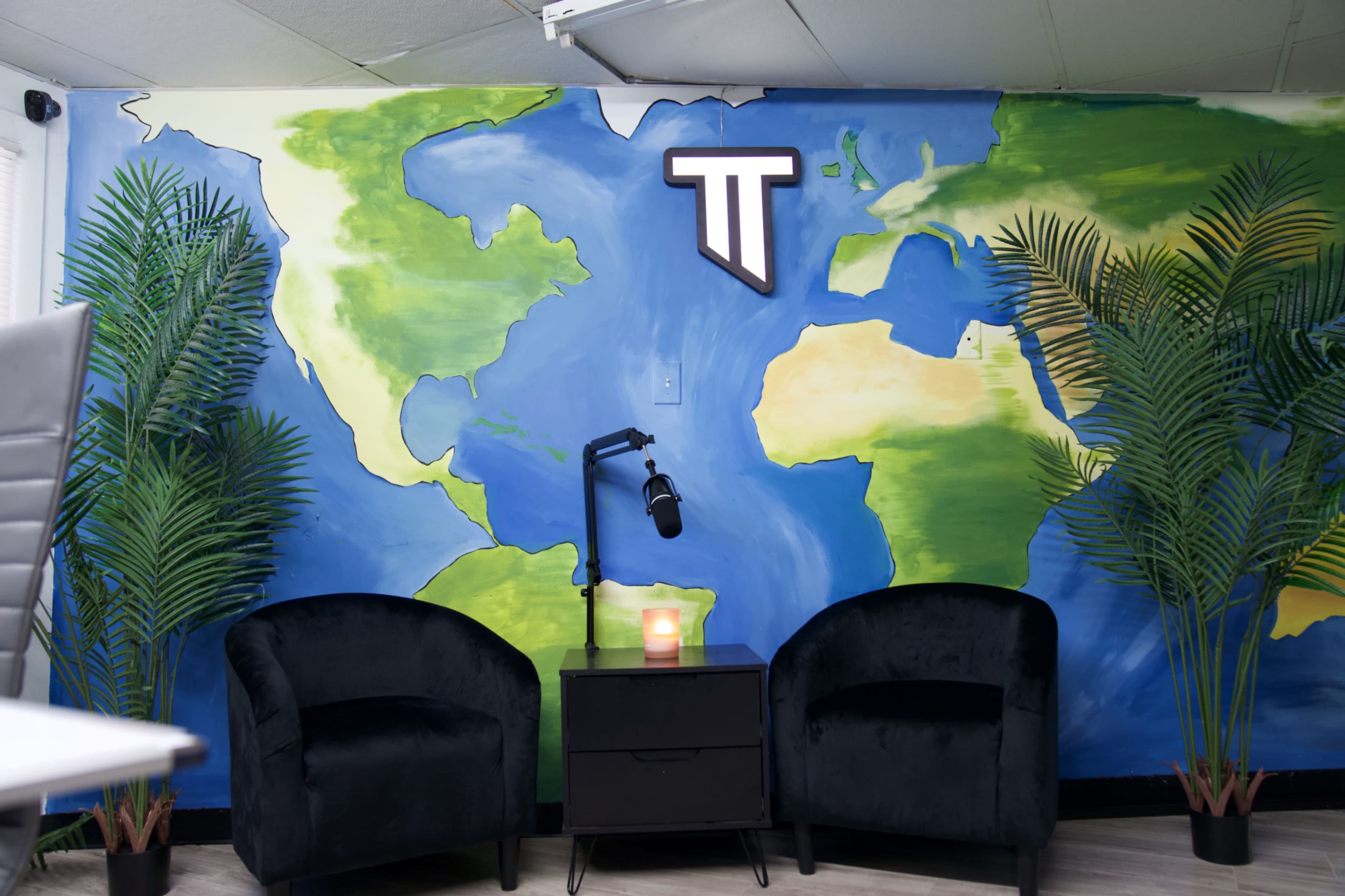 The image shows a wall mural of a world map with two black chairs and a small table beneath a microphone.