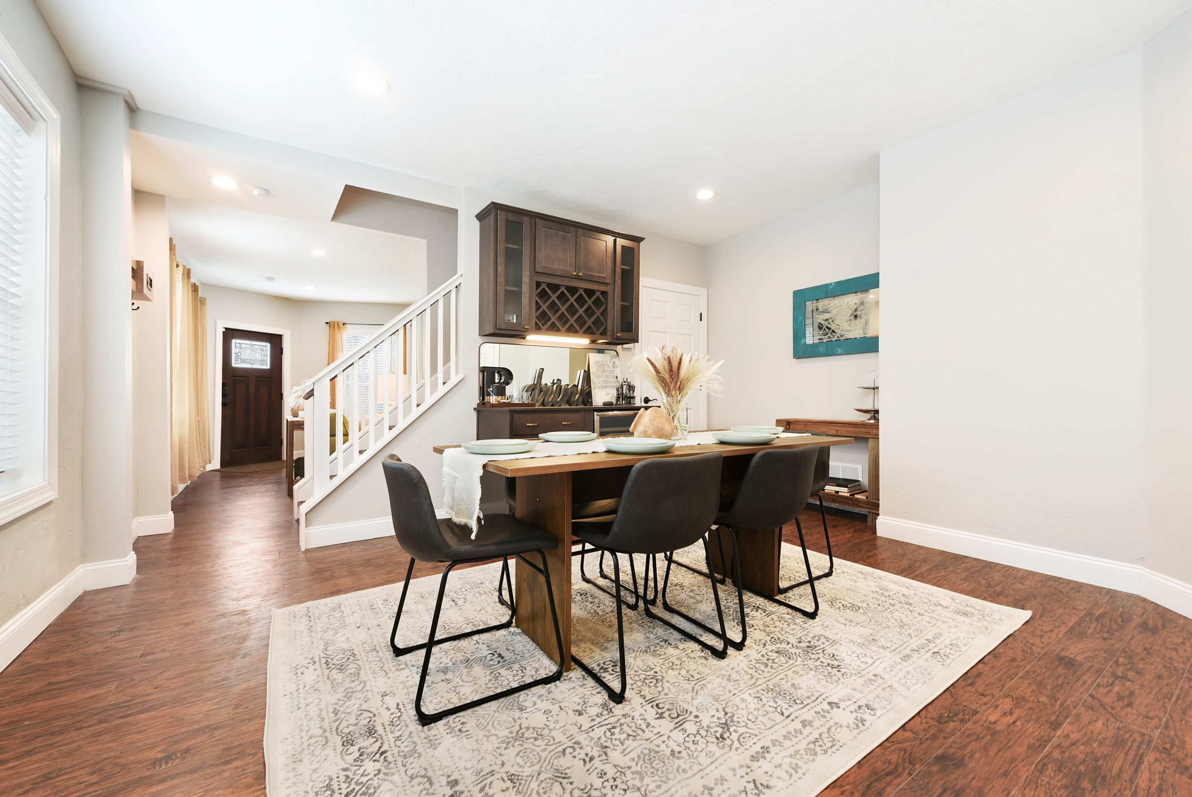 A dining area features a wooden table with chairs on a patterned rug, bordered by a staircase and kitchen.