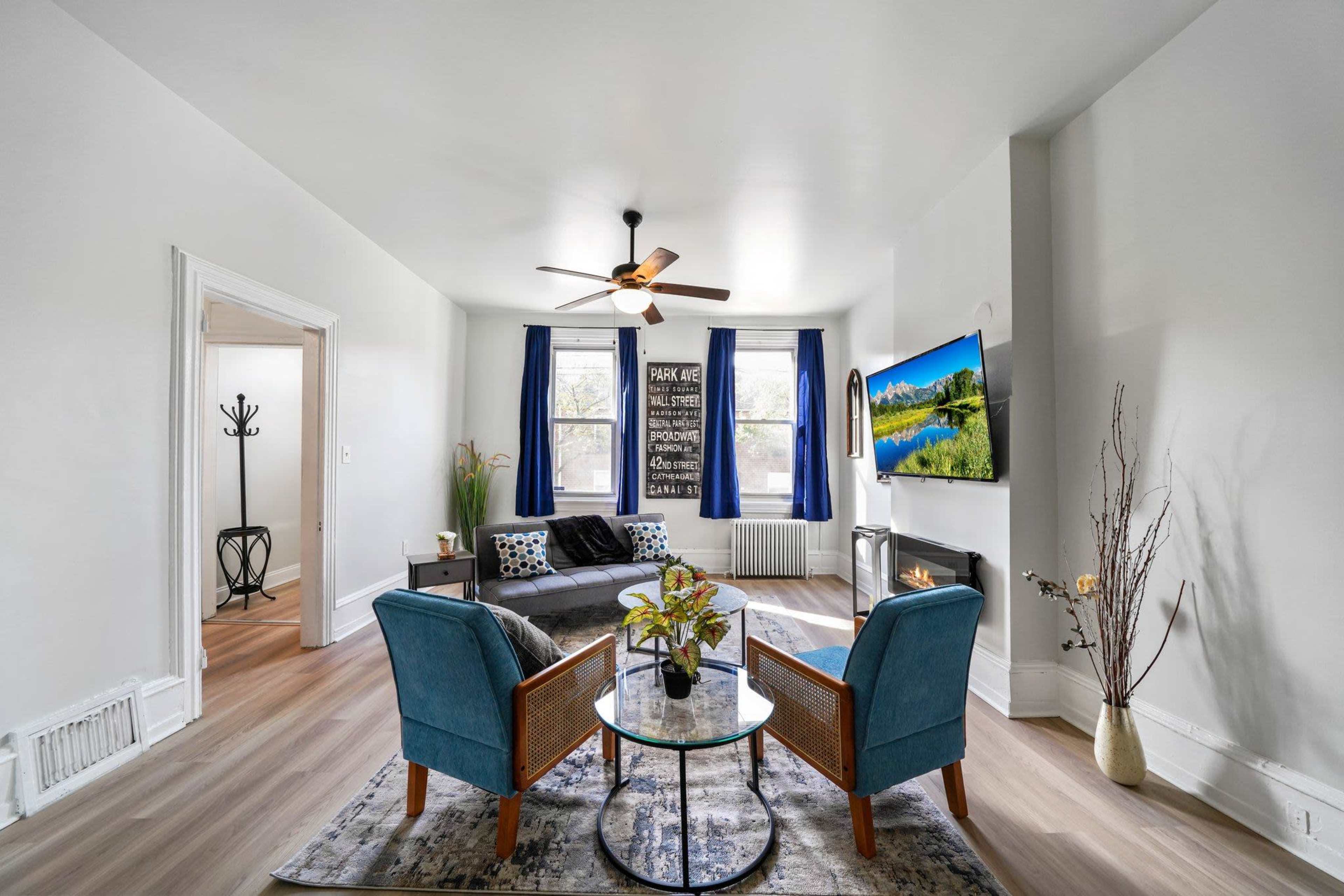 A modern living room with two blue chairs facing a circular glass table, a sofa, and a wall-mounted TV, all illuminated by natural light from large windows.