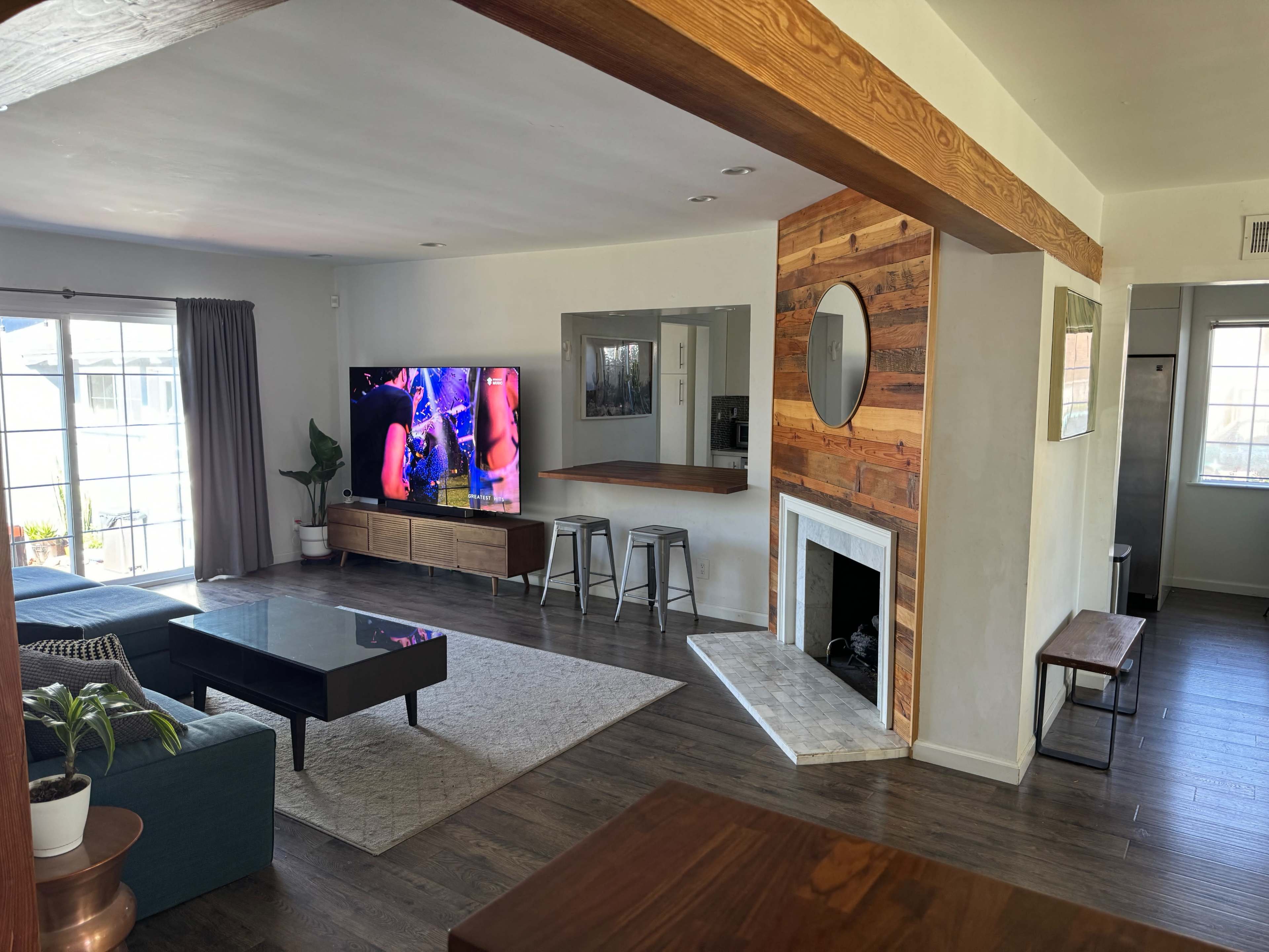 A modern living room with a gray couch, a large television mounted on the wall, and a rustic wooden accent feature surrounding a circular mirror and fireplace.