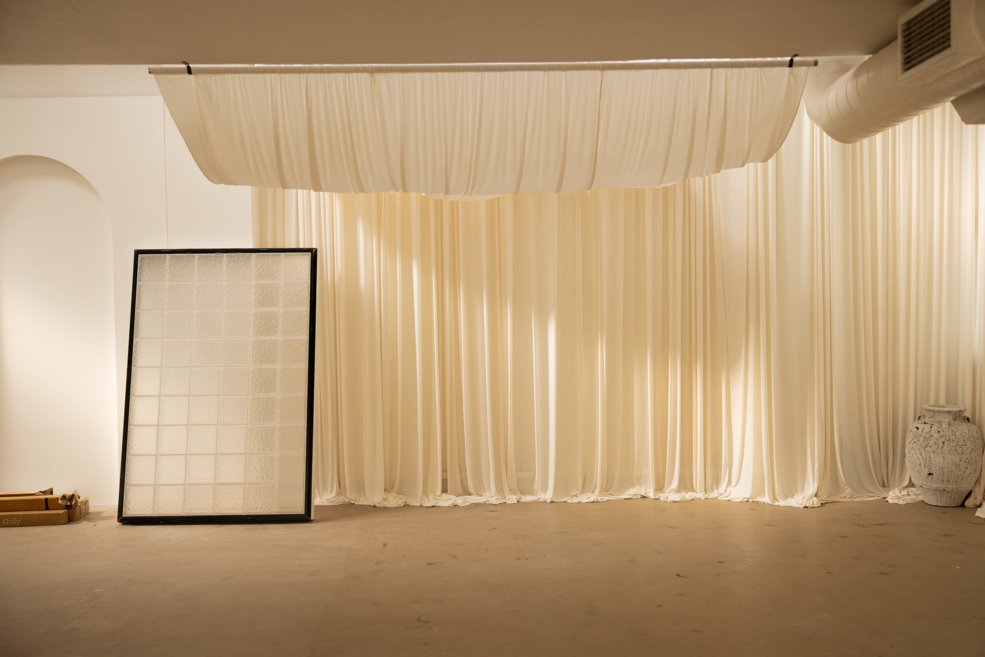 The image shows a spacious room featuring a large white curtain backdrop, a black-framed grid panel, and minimal furnishings on the floor.