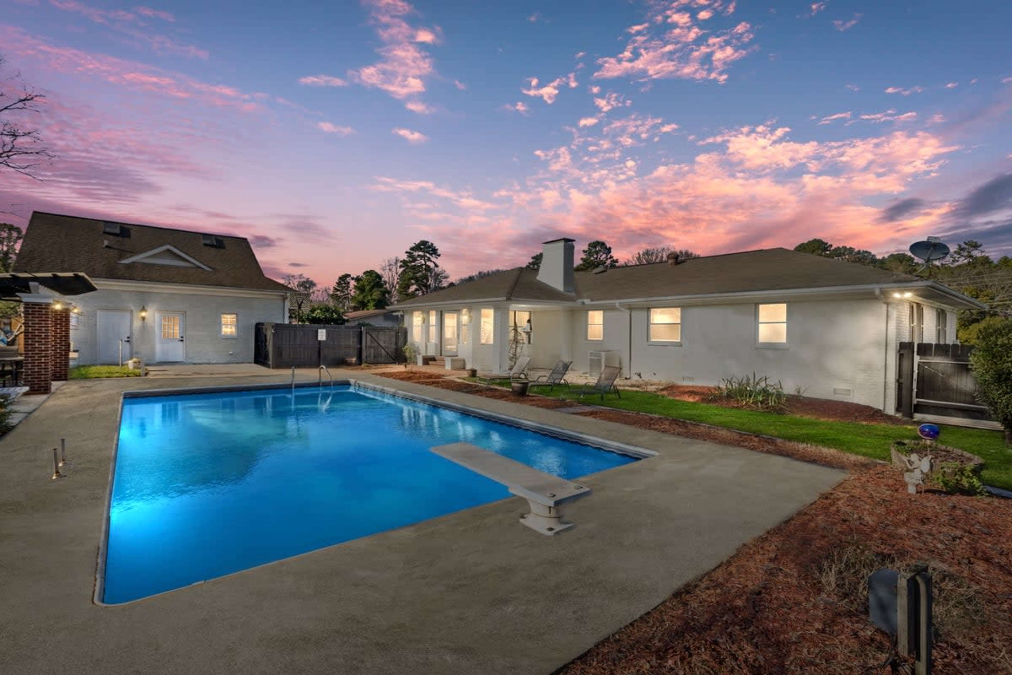 A rectangular swimming pool with a diving board is situated in a backyard, surrounded by grass and flower beds, under a colorful sky at sunset.