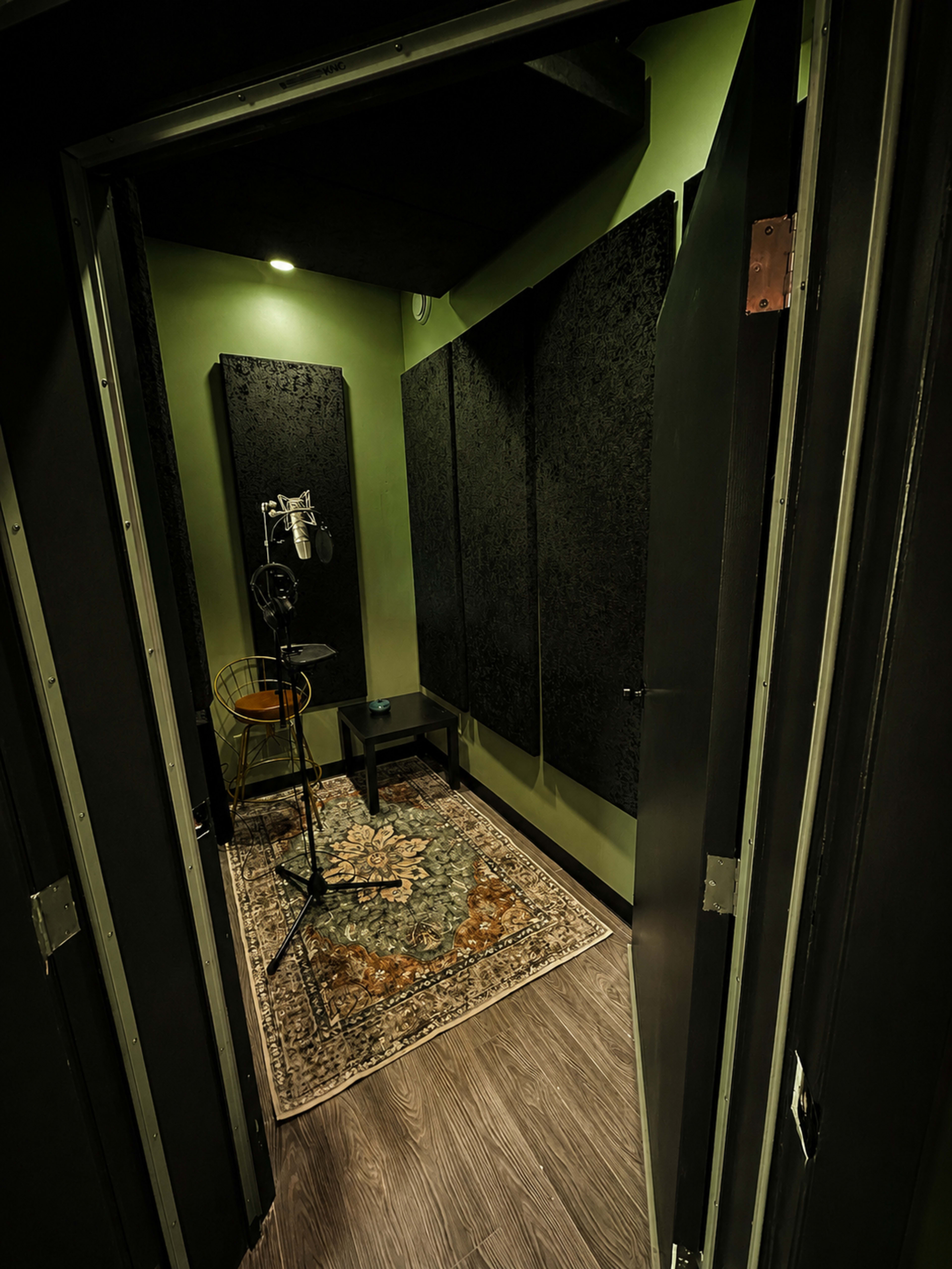 The image shows the interior of a soundproof recording booth with acoustic panels on the walls, a microphone on a stand, and a small table inside.