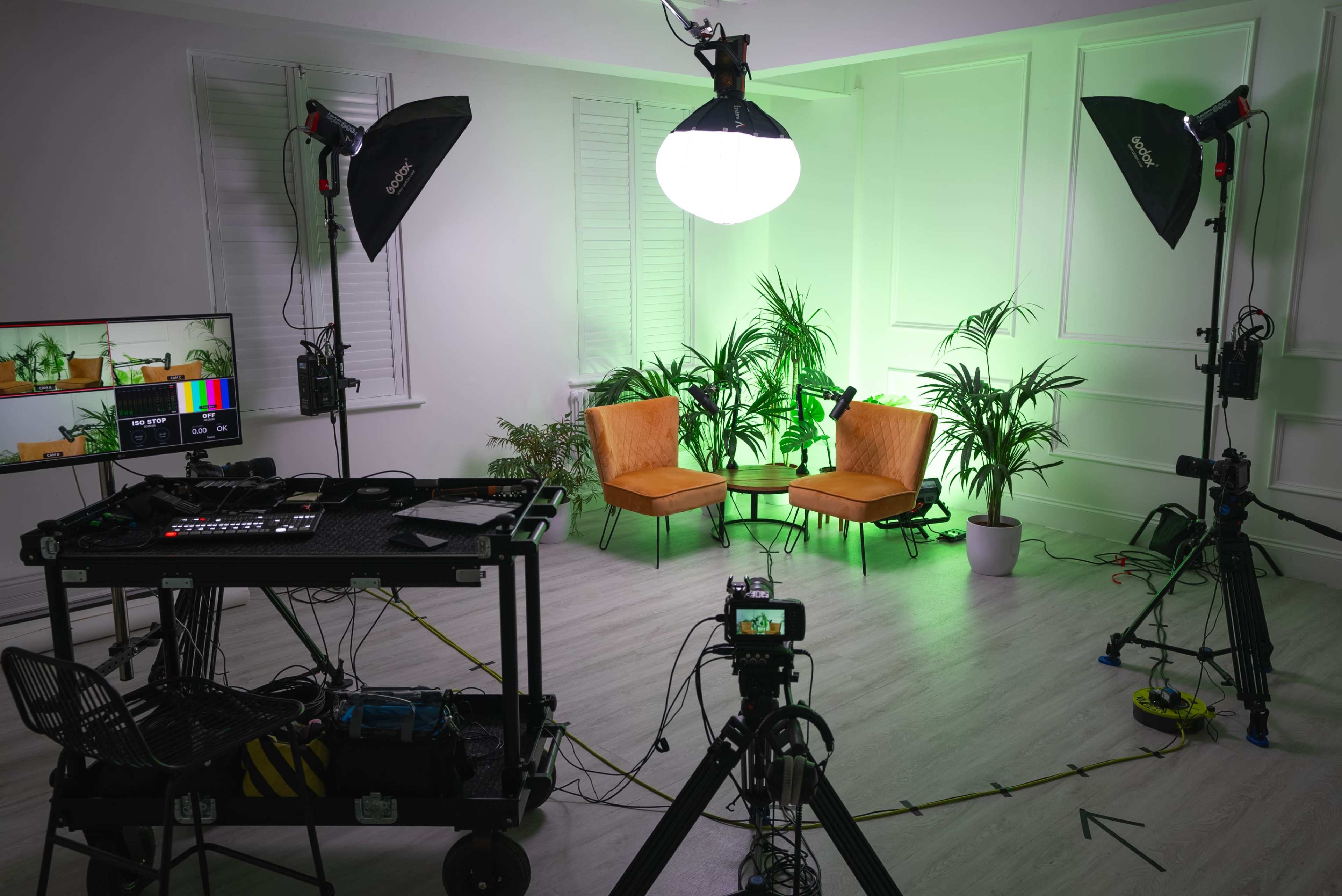 A well-equipped photo studio features two orange chairs, bright lighting, and various pieces of filming equipment arranged around the space.
