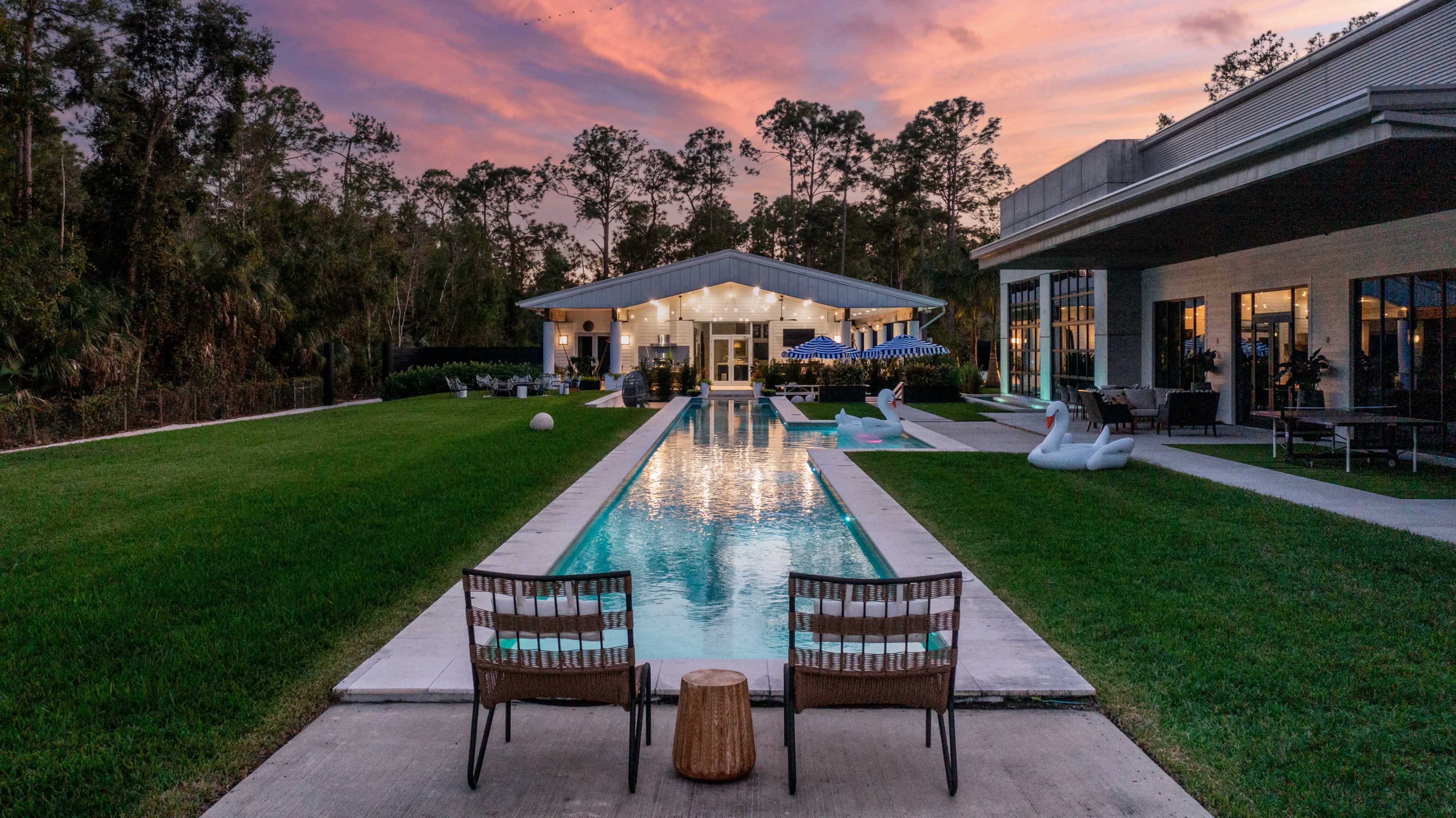A modern home with a swimming pool surrounded by lush grass, featuring two chairs and a wooden stool in the foreground against a sunset sky.