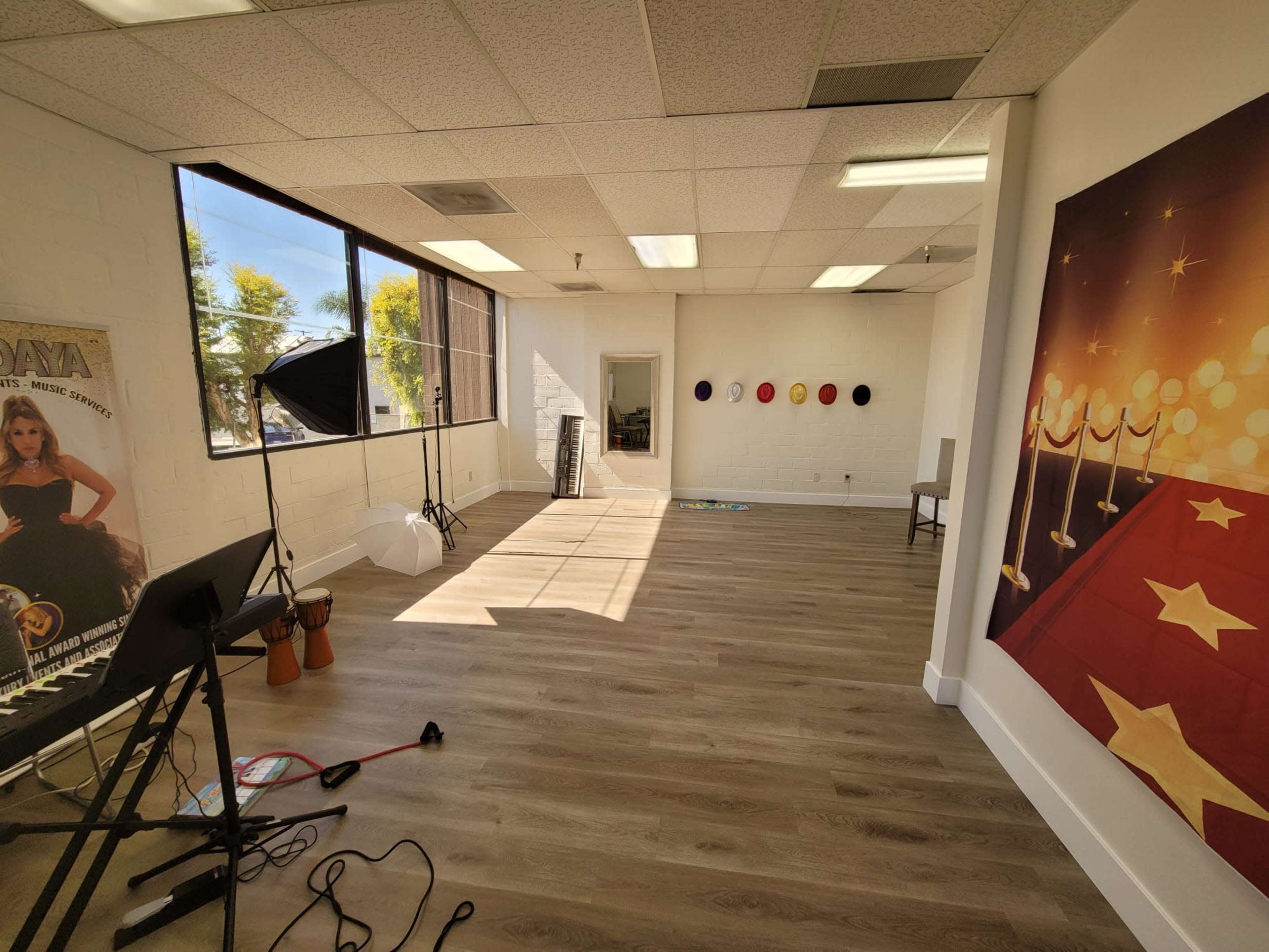 An empty studio space features wooden flooring, large windows letting in natural light, and a backdrop of a red carpet with spotlights on one wall.