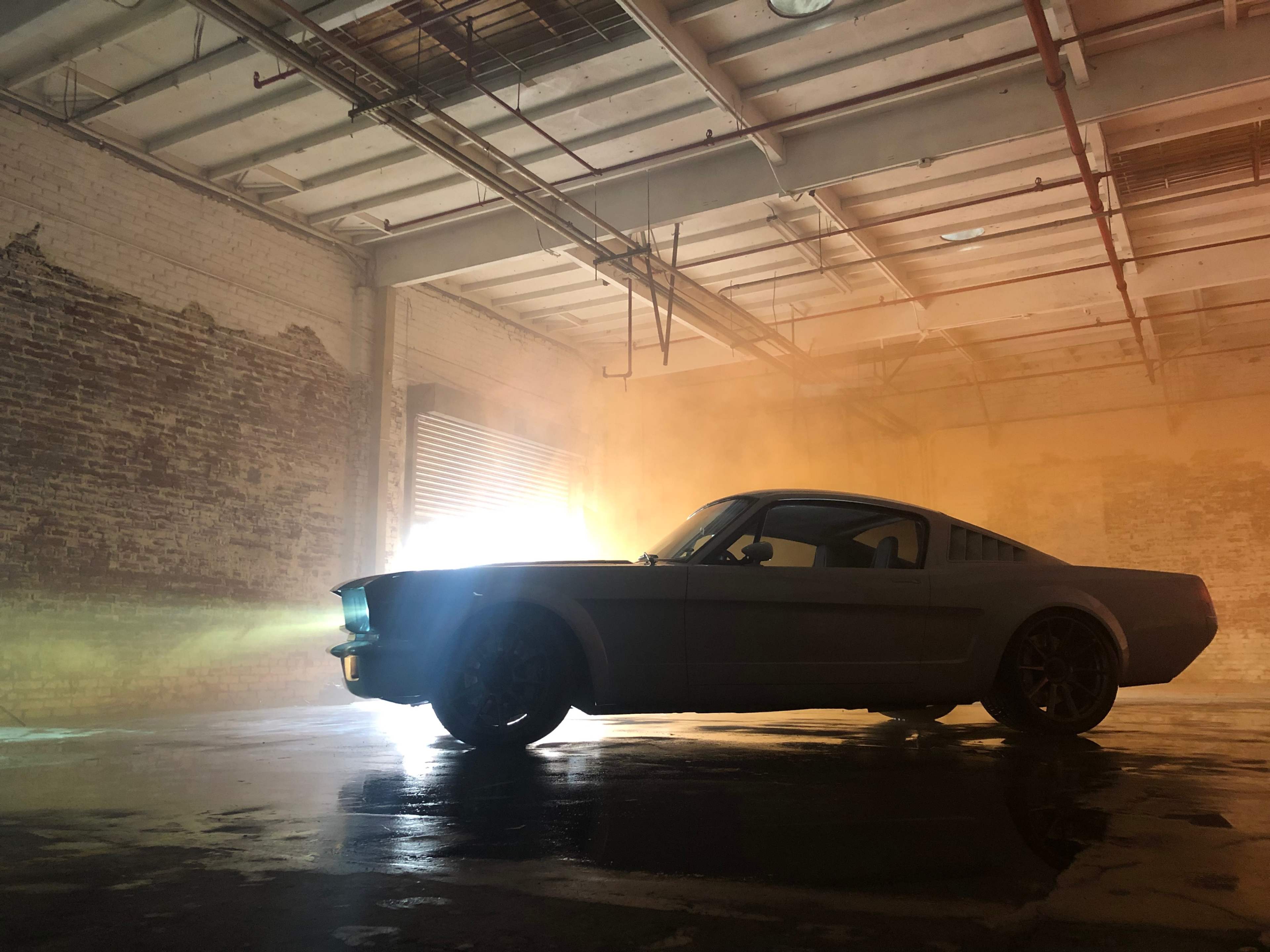 A classic car is silhouetted against a backdrop of smoke and light in an industrial space.