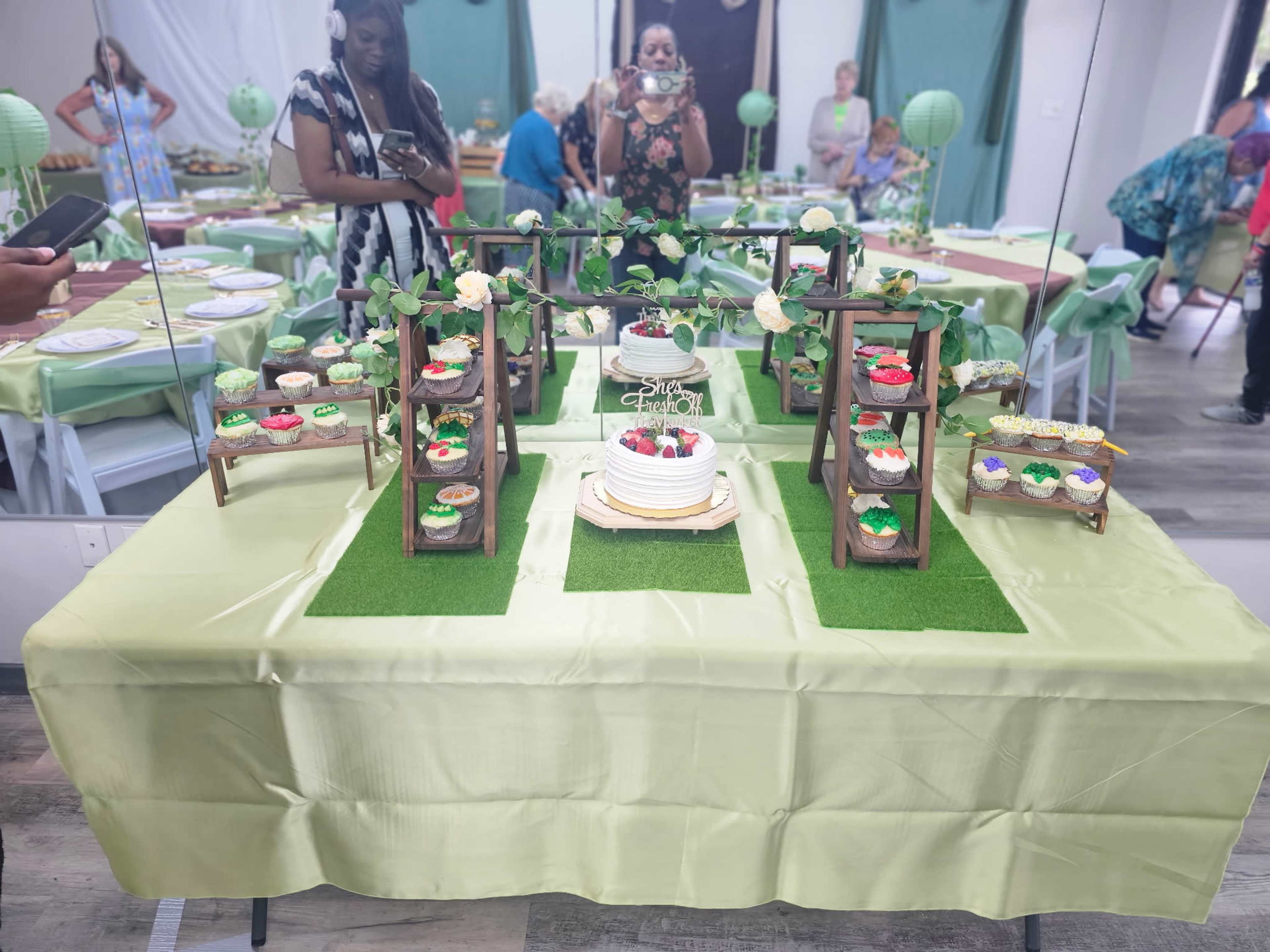 A decorated table featuring a variety of colorful cakes and desserts arranged on tiered stands, with guests and a mirror reflecting the scene in the background.