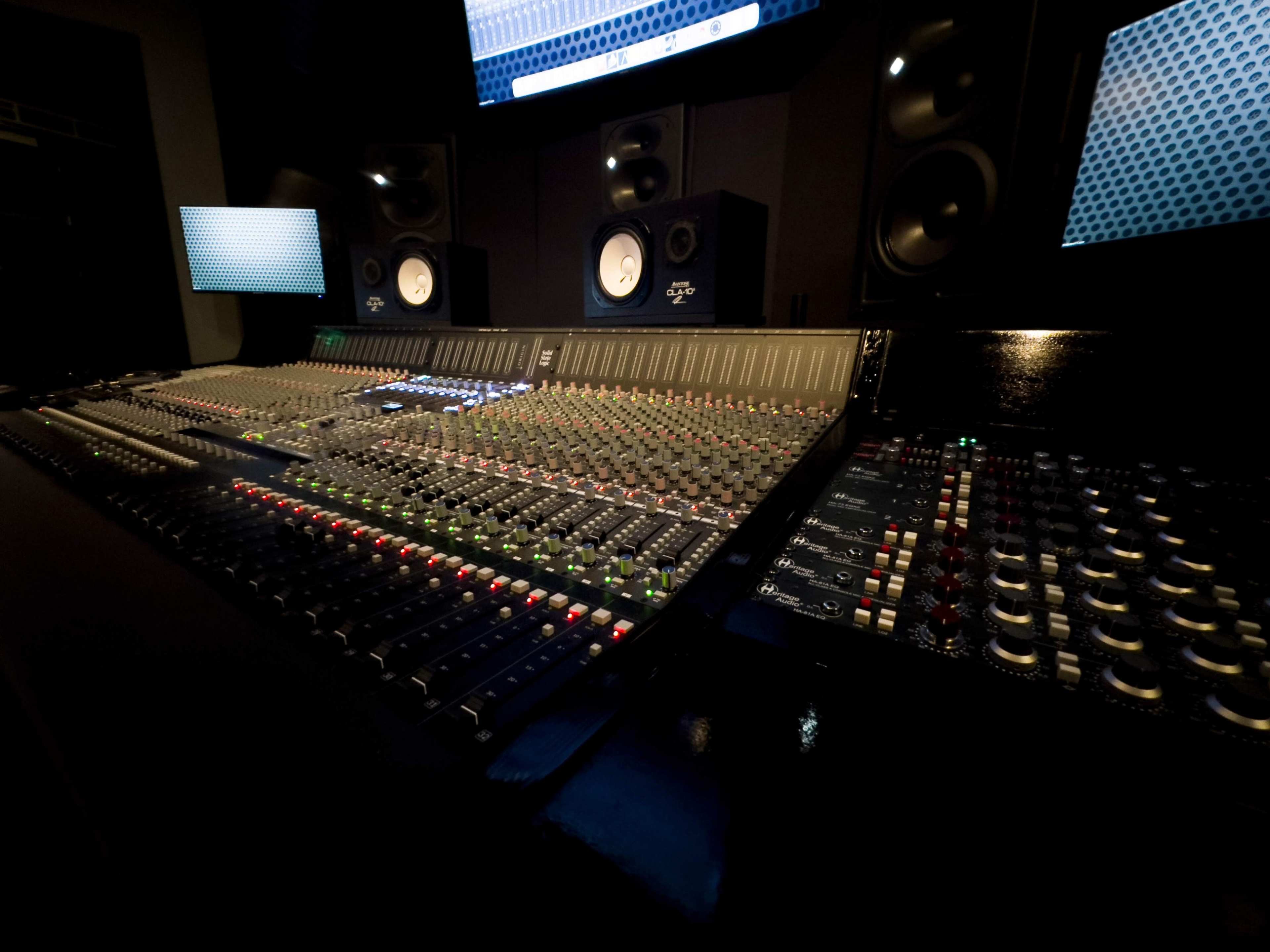 A music recording studio features a large mixing console surrounded by monitors and studio speakers.