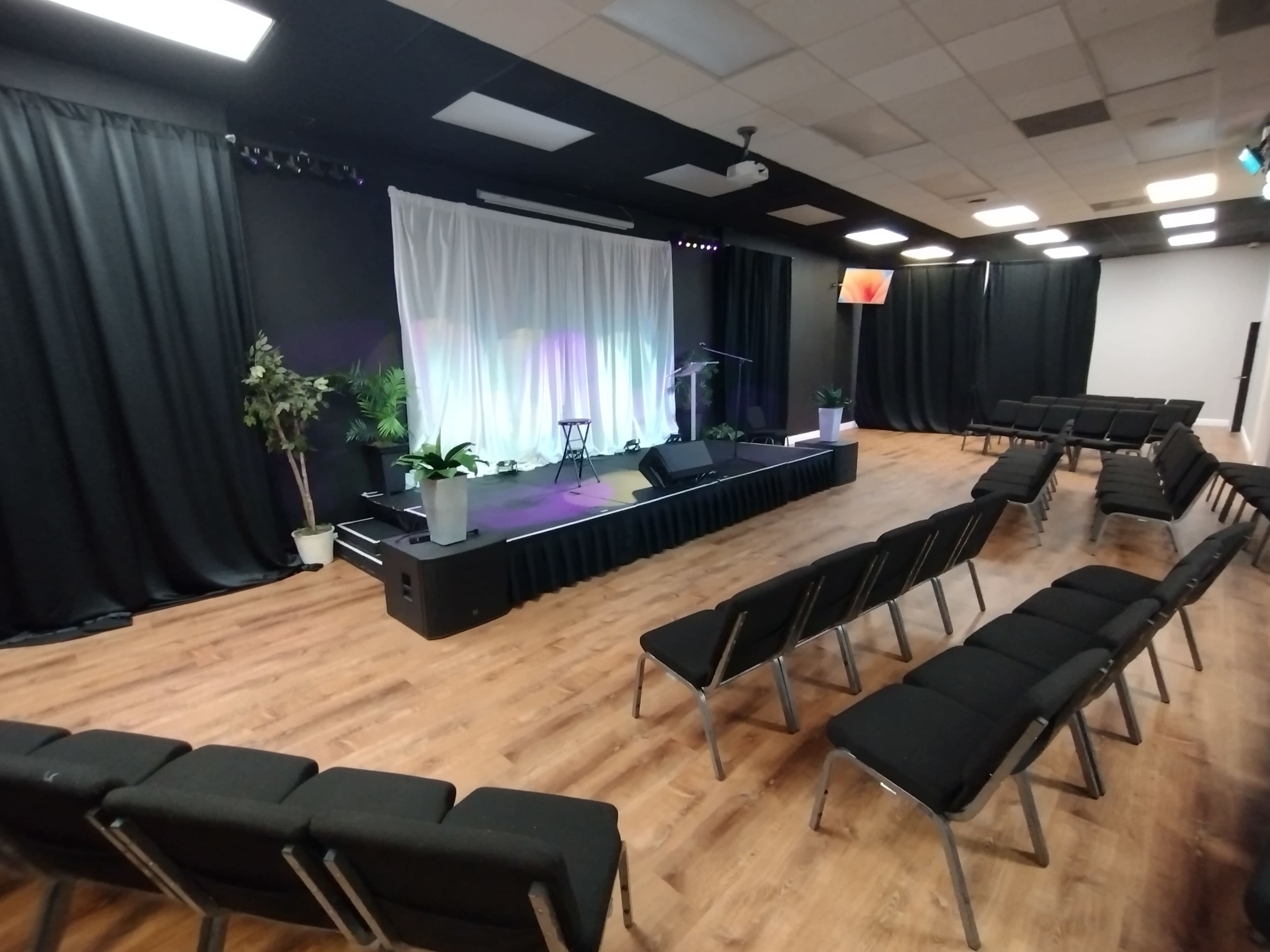 A stage with a white backdrop and plants, surrounded by rows of black chairs in a large room with wooden flooring.