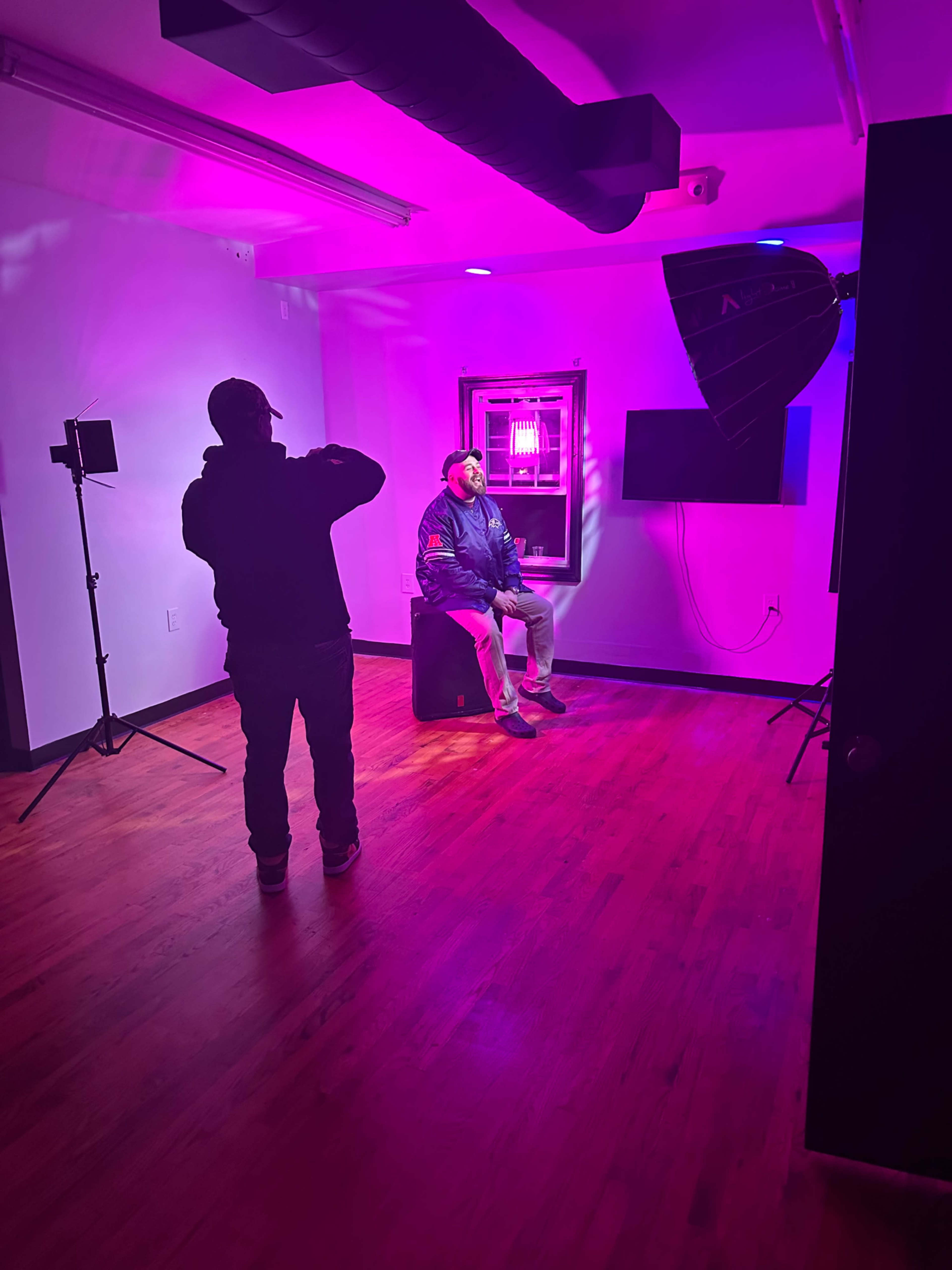 A person sits on a stool in a brightly lit studio with purple lighting, while another person photographs them.