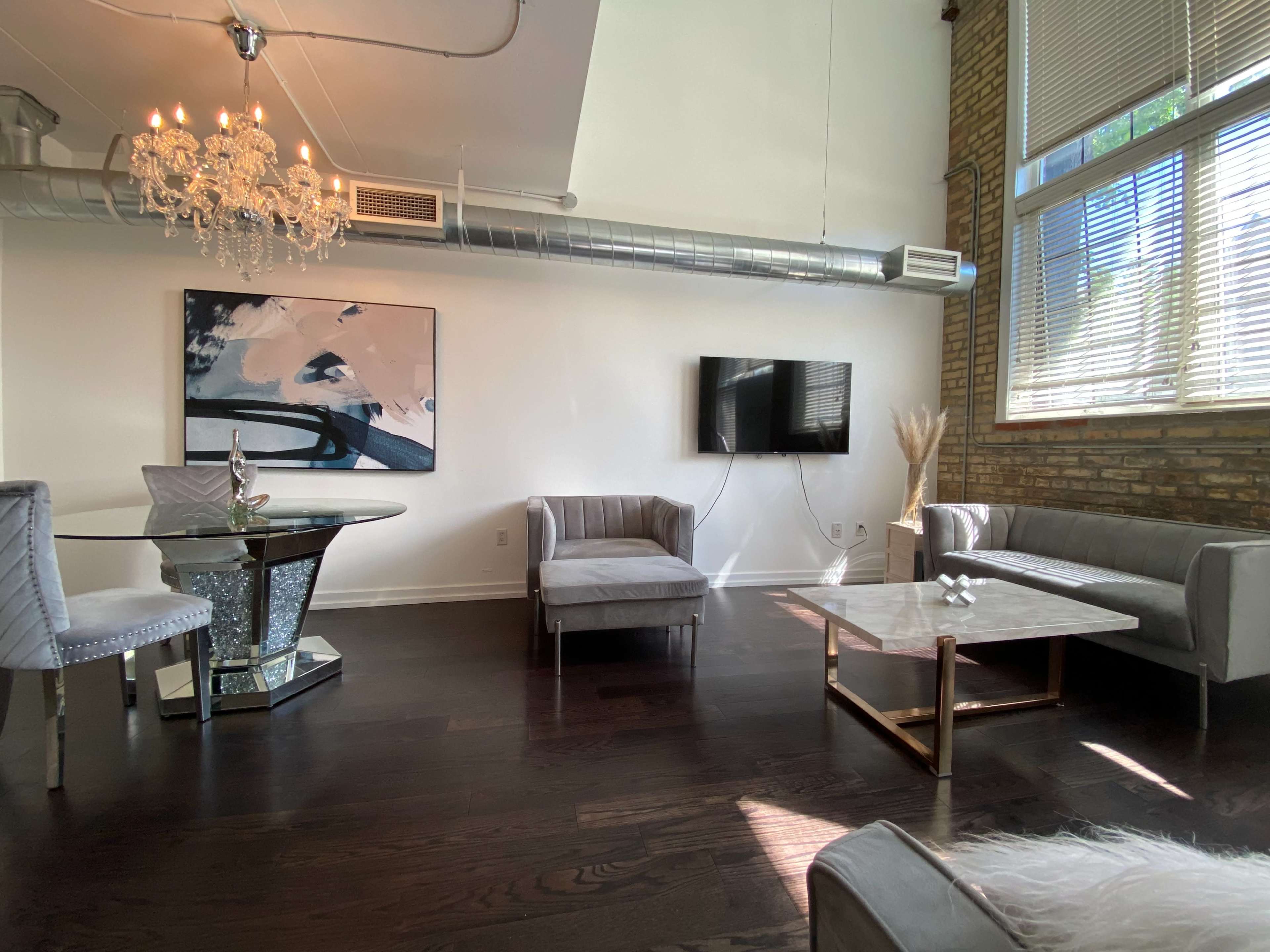 The scene shows a modern living area with a round dining table, a gray sofa, a light gray armchair, a coffee table, a flat-screen TV, and a chandelier.