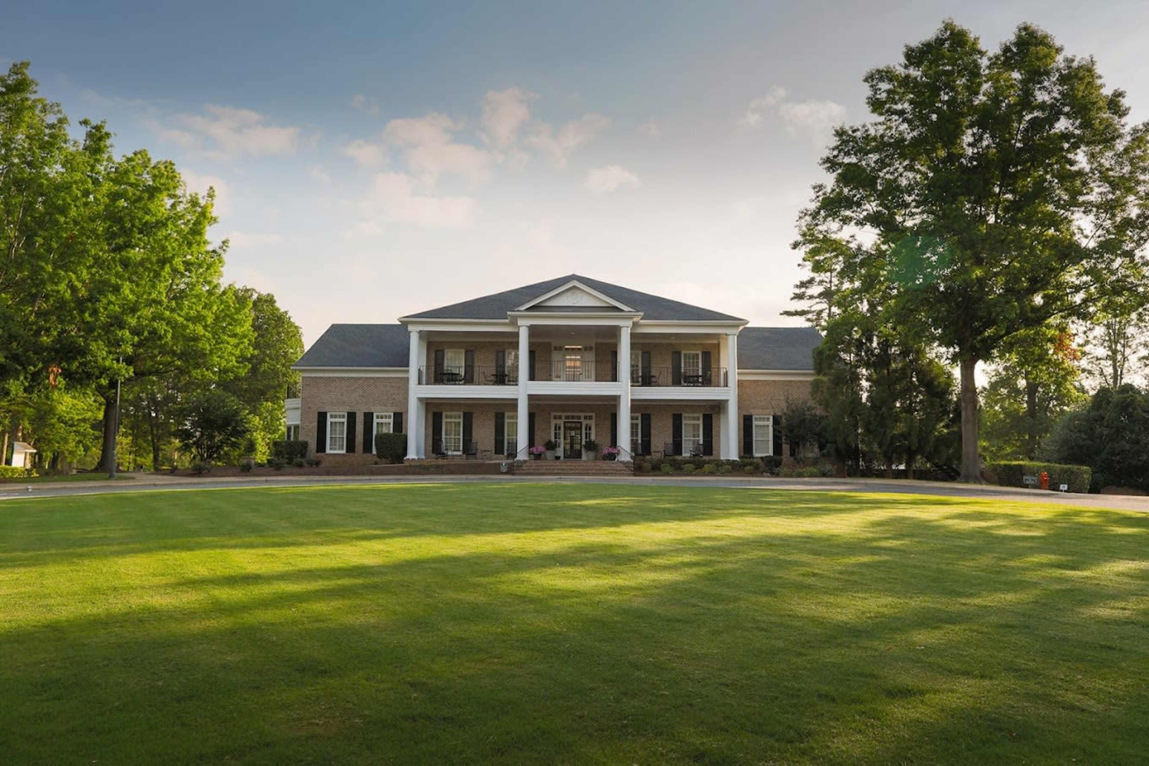 A large two-story brick house with a well-manicured lawn and several trees in the foreground is depicted.
