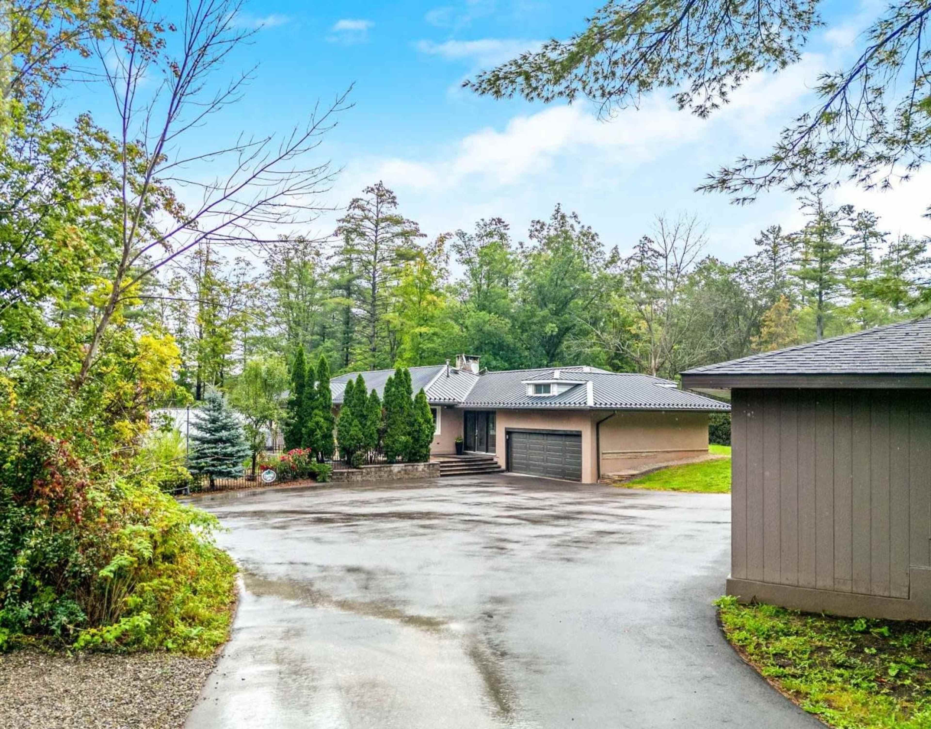A modern house with a gray exterior is surrounded by trees, sitting at the end of a gravel driveway.