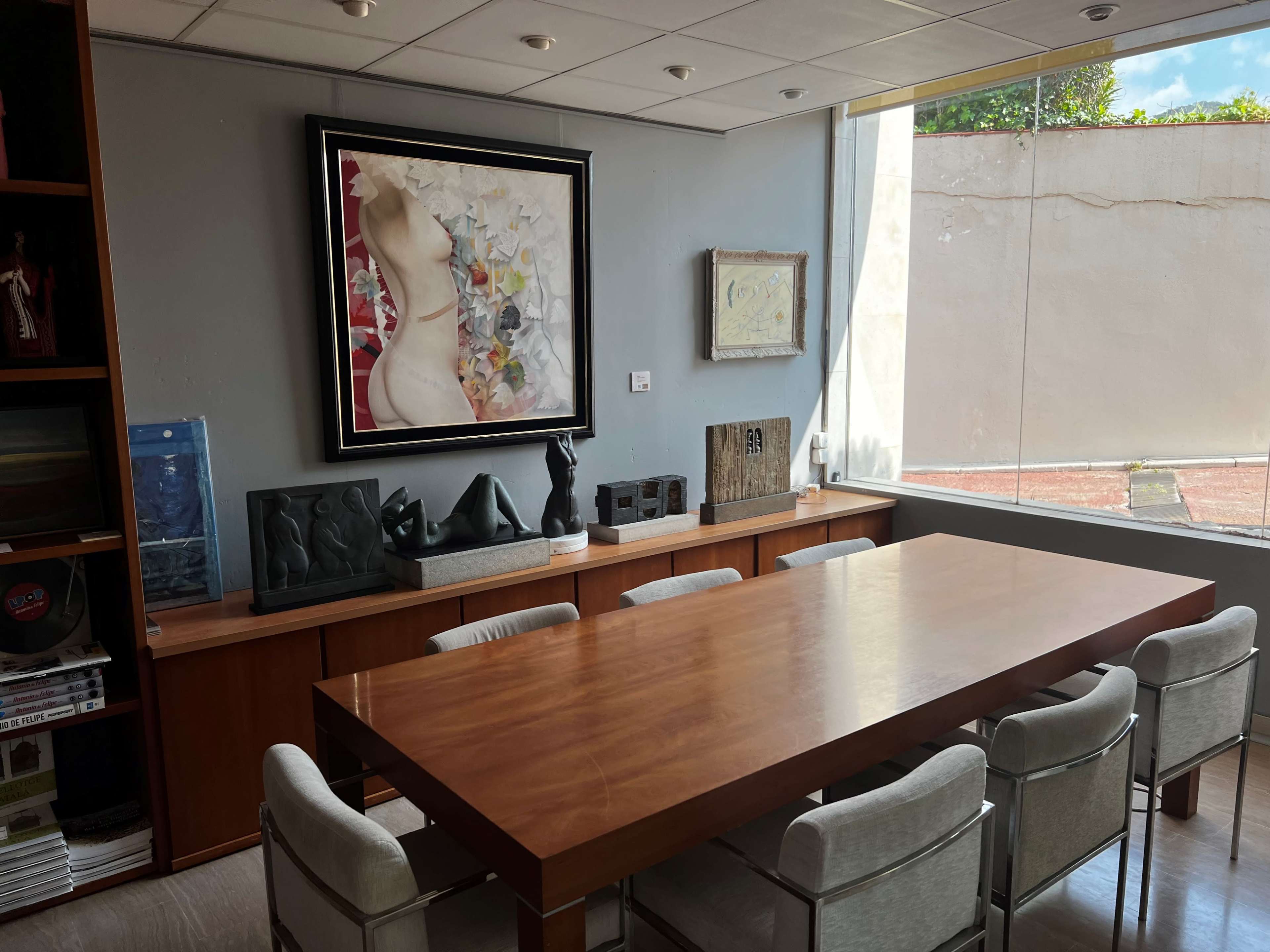 The image shows a modern conference room with a large wooden table surrounded by eight chairs, artwork on the walls, and natural light coming through a window.