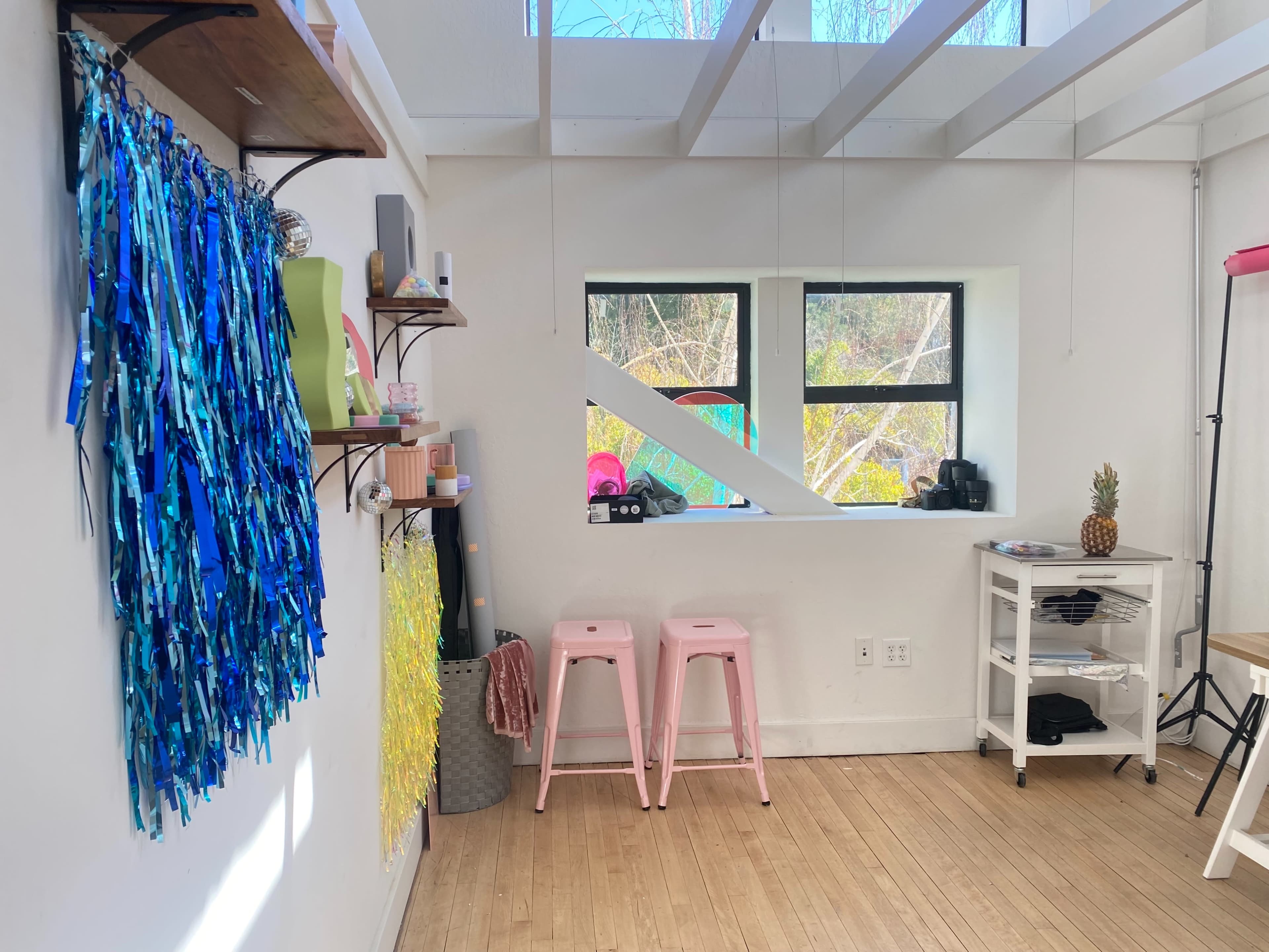 The image shows a bright, minimalistic room with colorful decorations on one wall, two pink stools, and a workspace featuring a table and storage cart.