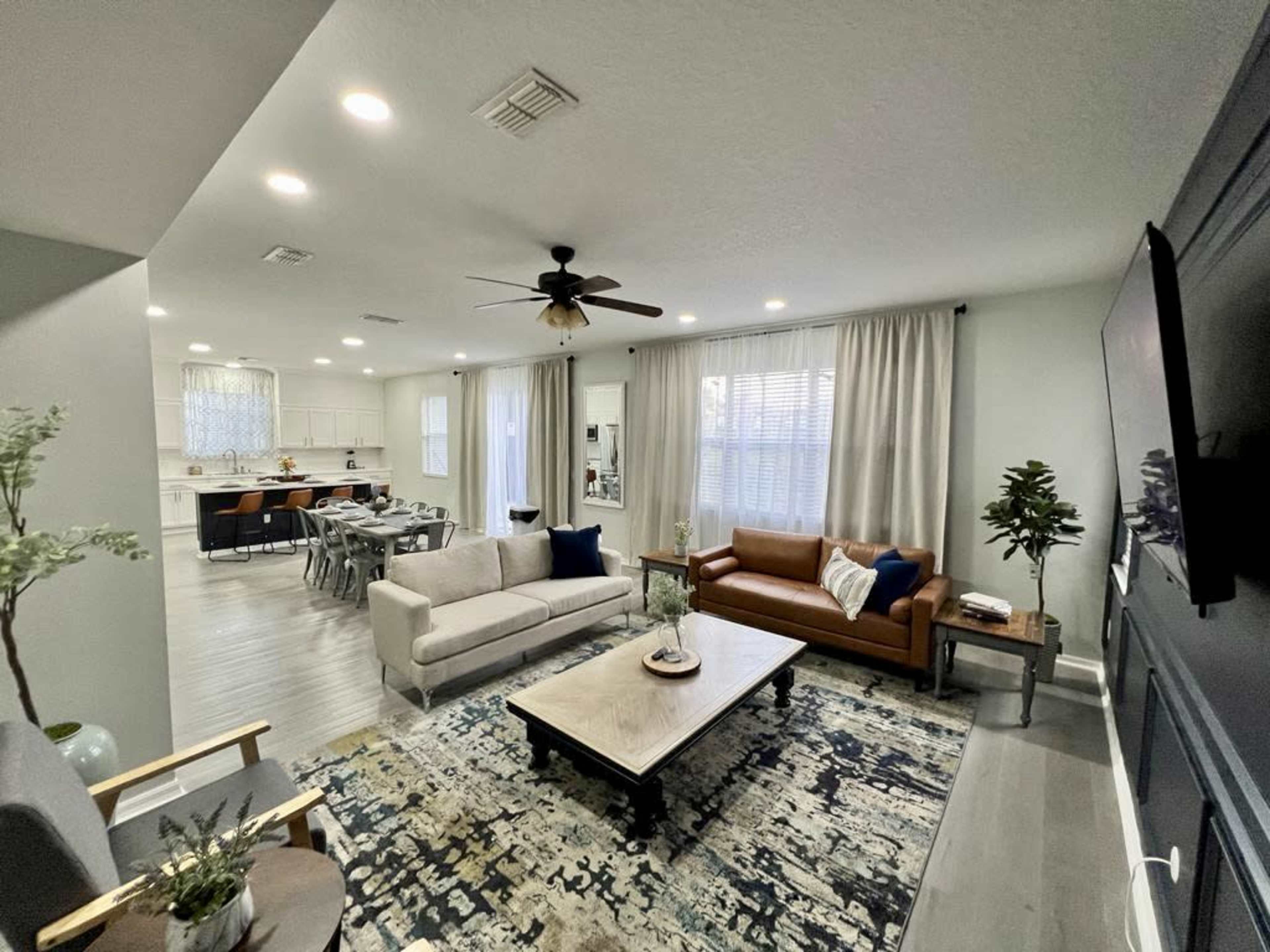 The image shows a modern living room with a light-colored couch, a brown leather sofa, a coffee table, and a view of an open kitchen area in the background.