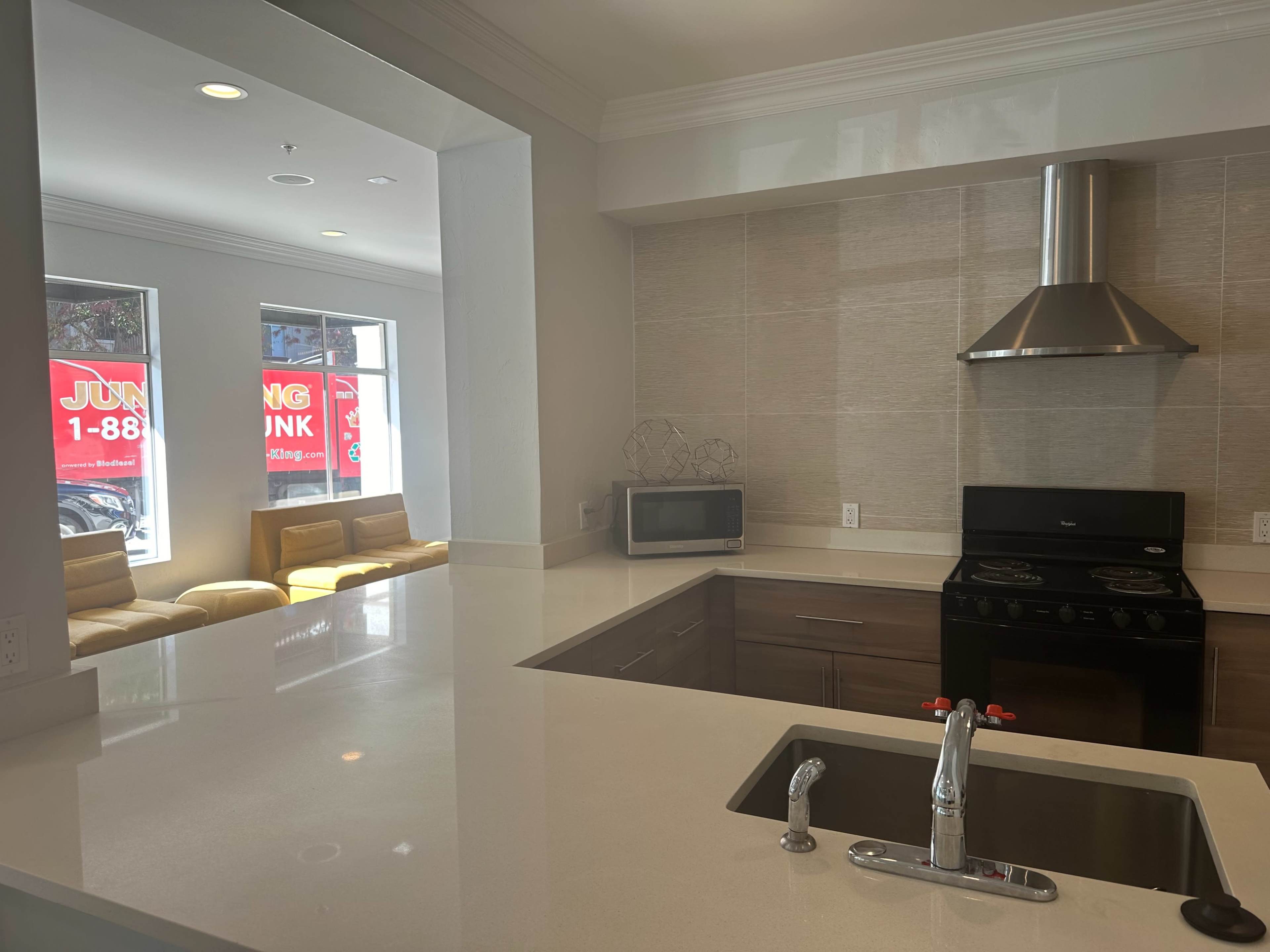 A modern kitchen with a stove, microwave, and a large countertop, overlooking a seating area with yellow chairs and windows.