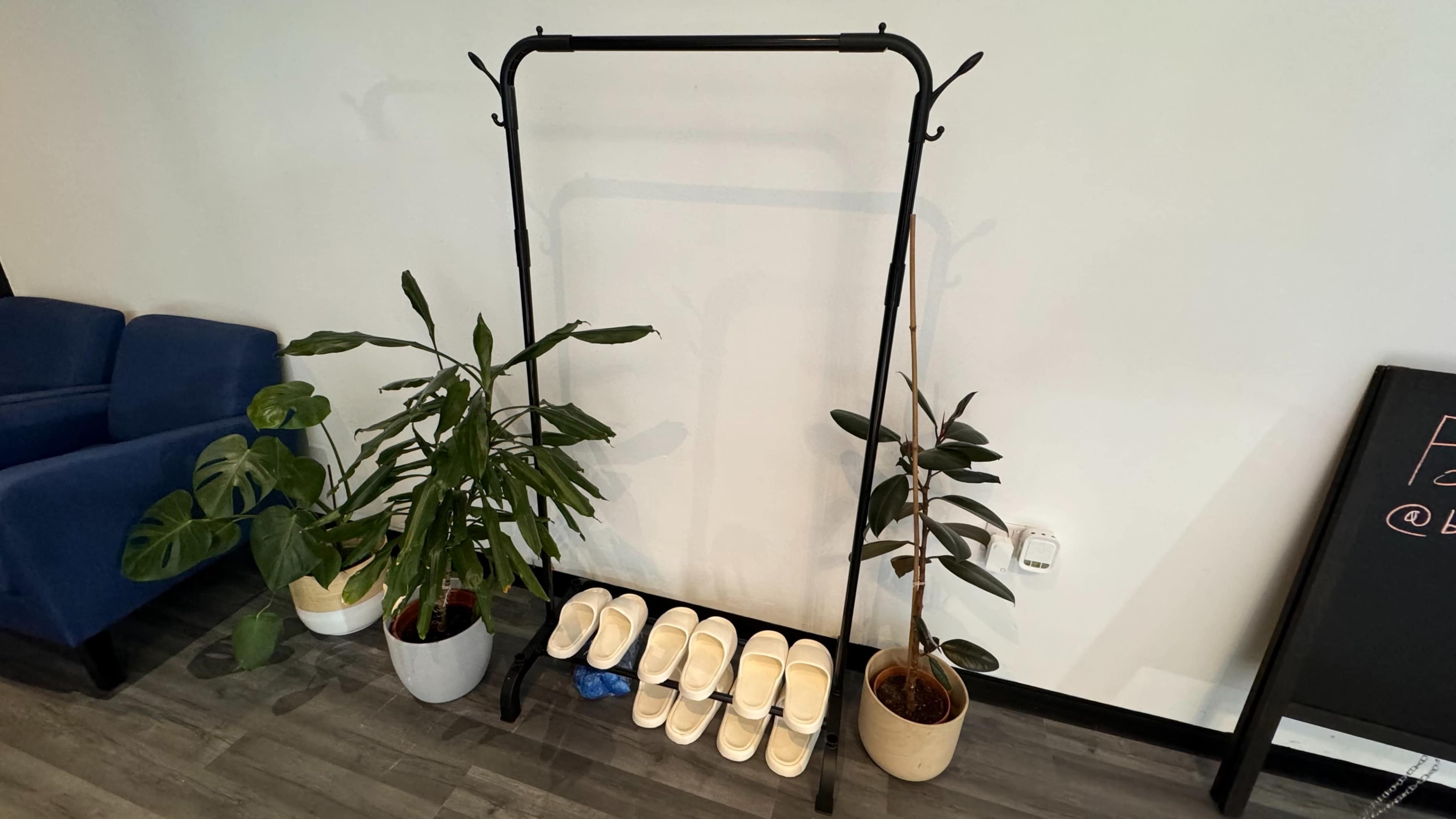A clothing rack stands against a wall next to two potted plants and a row of white slippers on the floor.
