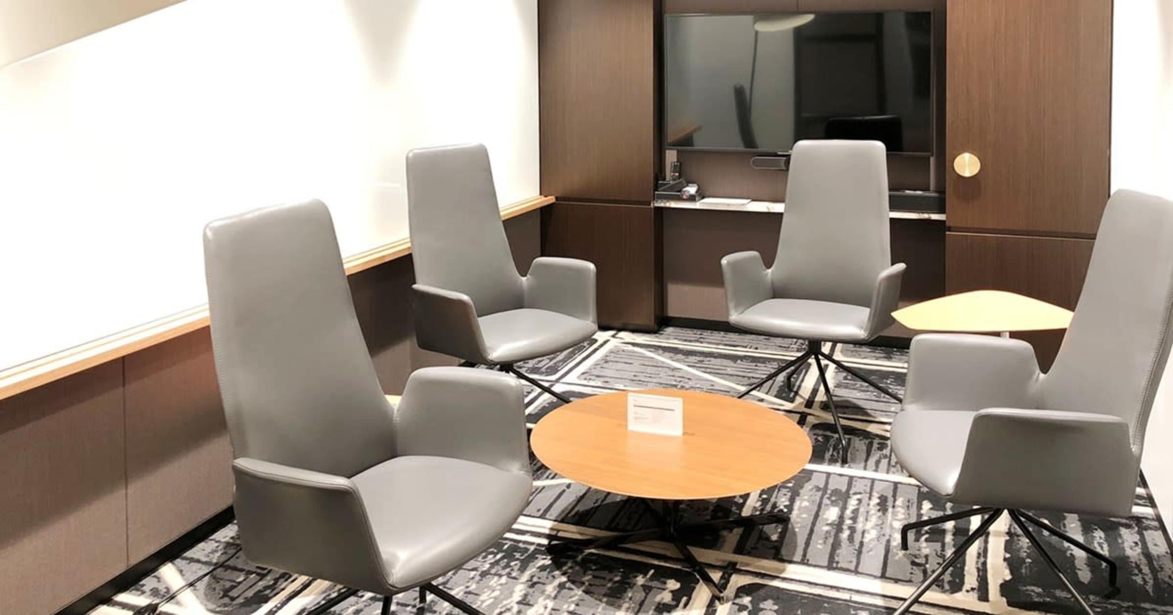 The image shows a small meeting room with four gray chairs arranged around a round wooden table on a patterned carpet.