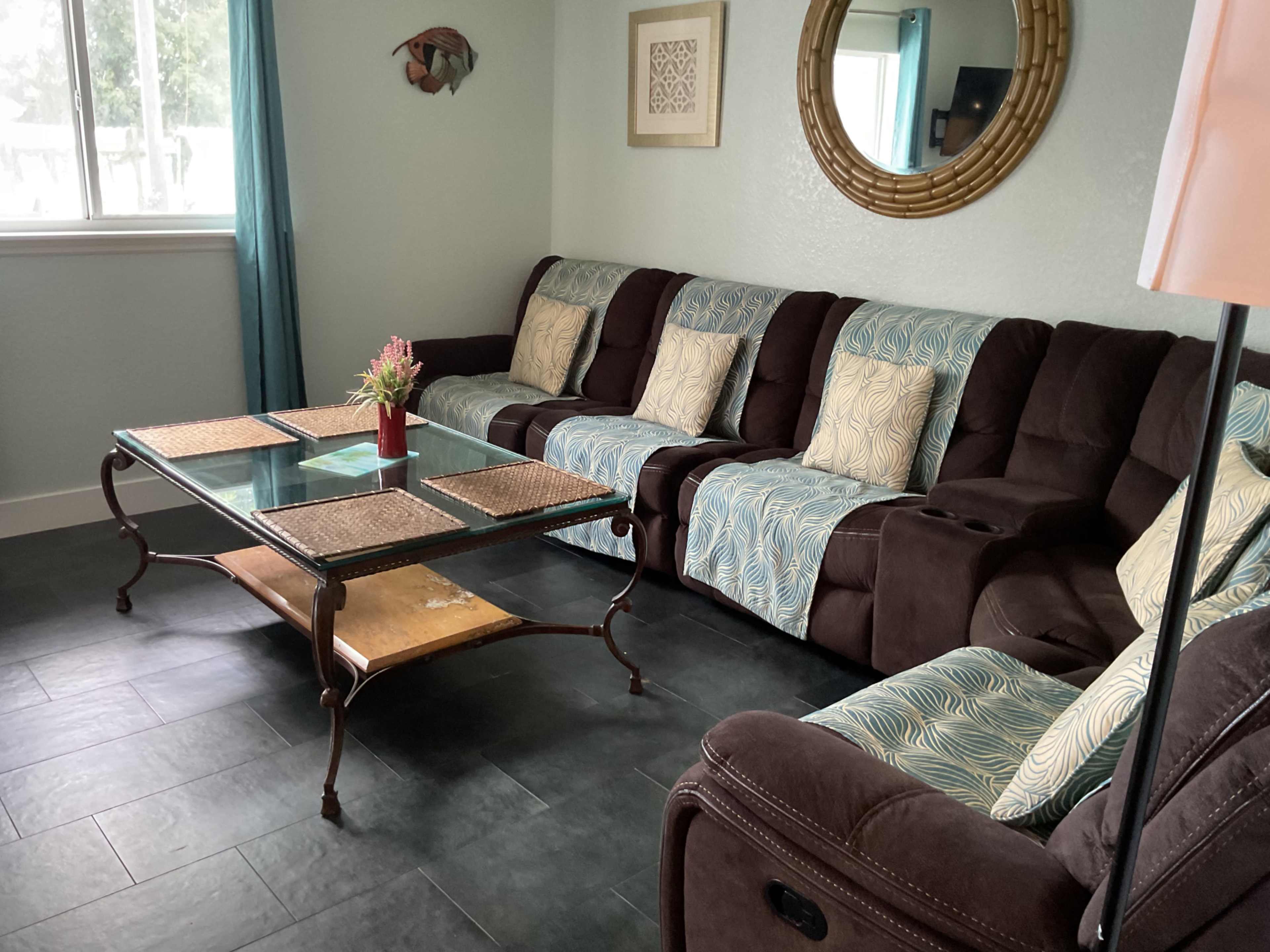 The image shows a living room with a sectional couch, a glass coffee table, and a large round mirror on the wall, all set against light blue walls.