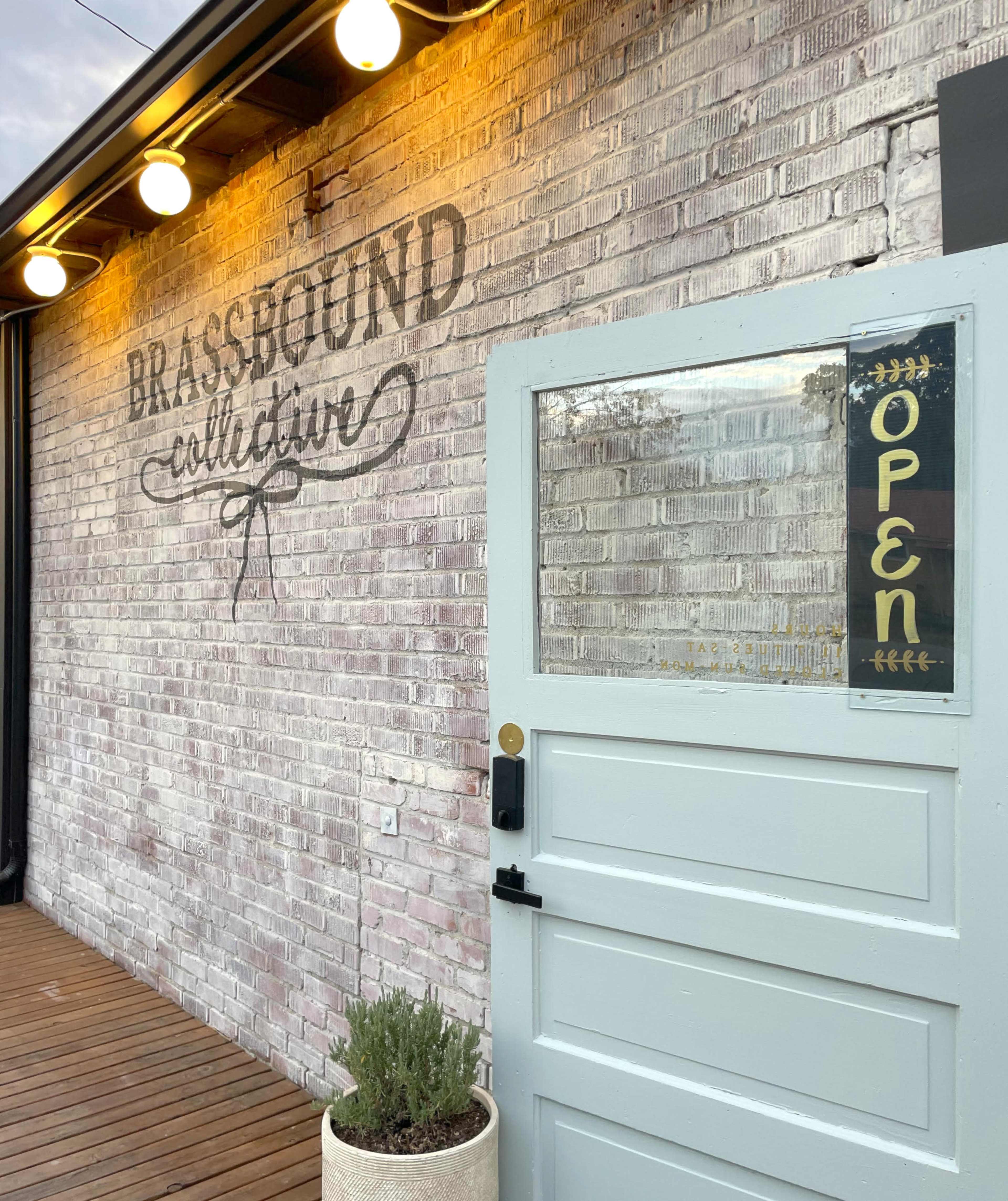 The image shows a light-colored door with an "OPEN" sign, set against a brick wall featuring the words "BRASSBOUND collection" and illuminated by string lights.