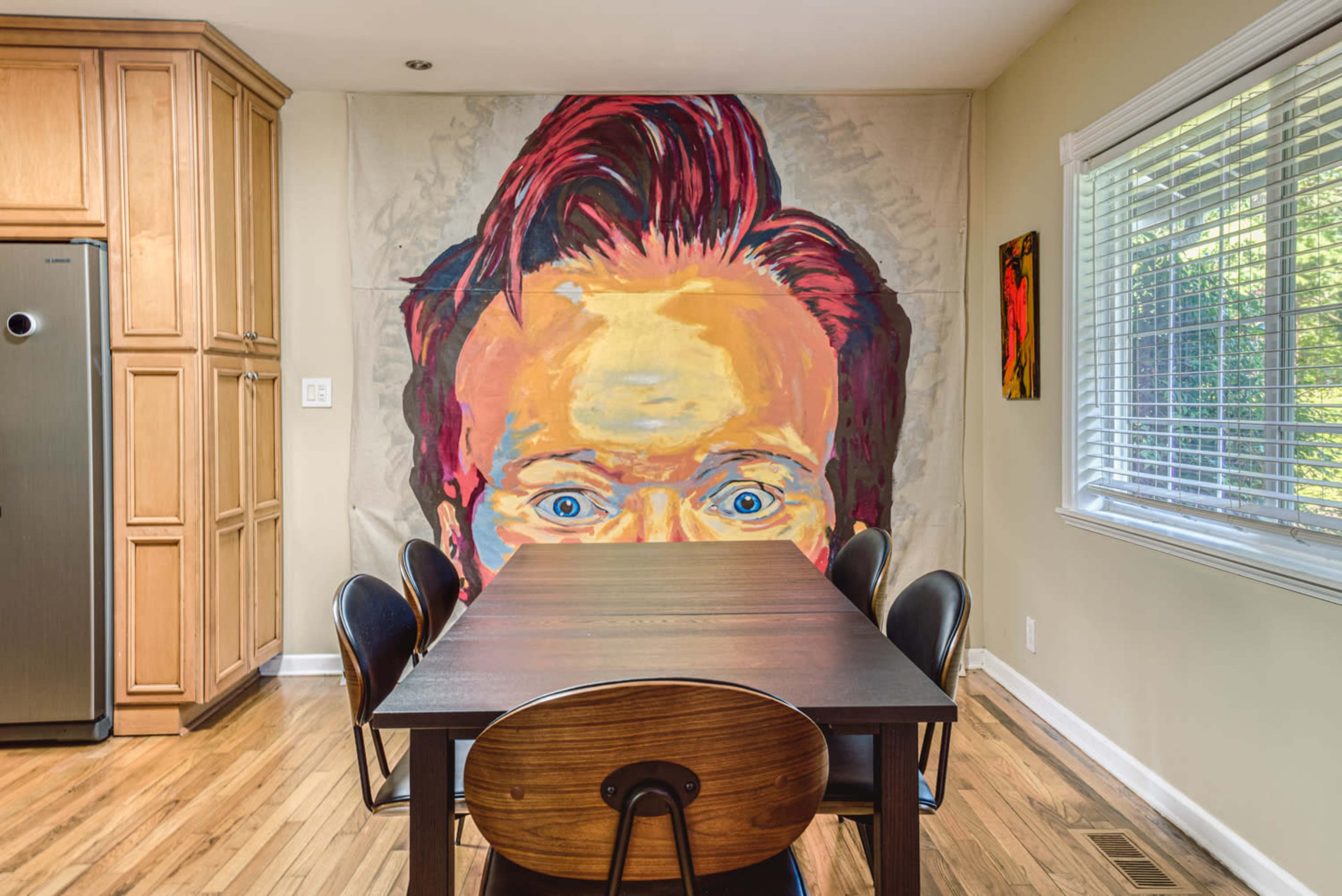 A large mural of a person's face dominates the wall above a modern dining table surrounded by black chairs in a well-lit kitchen.
