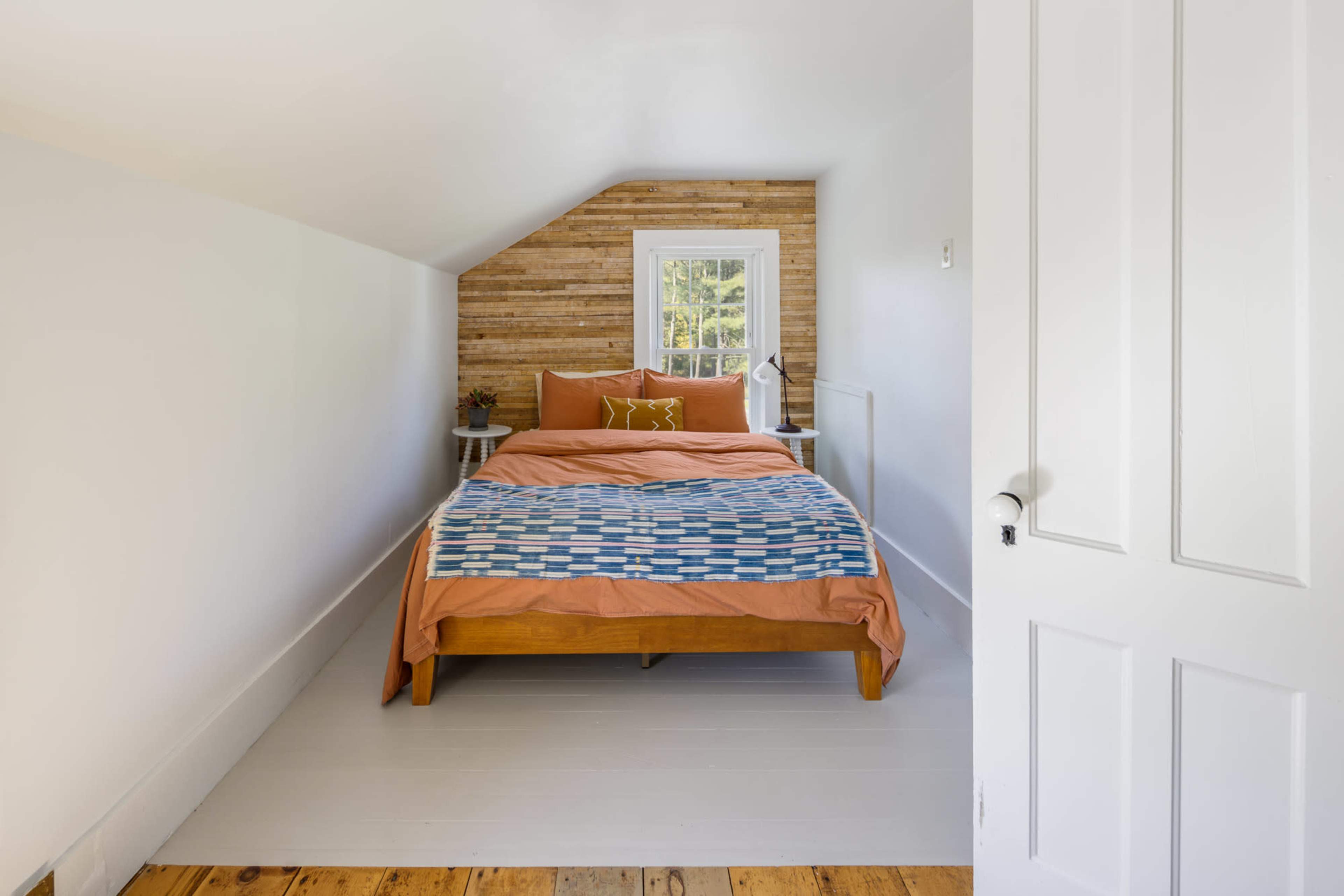 A small bedroom features a wooden bed with an orange and blue bedding set against a white wall with a wooden accent.