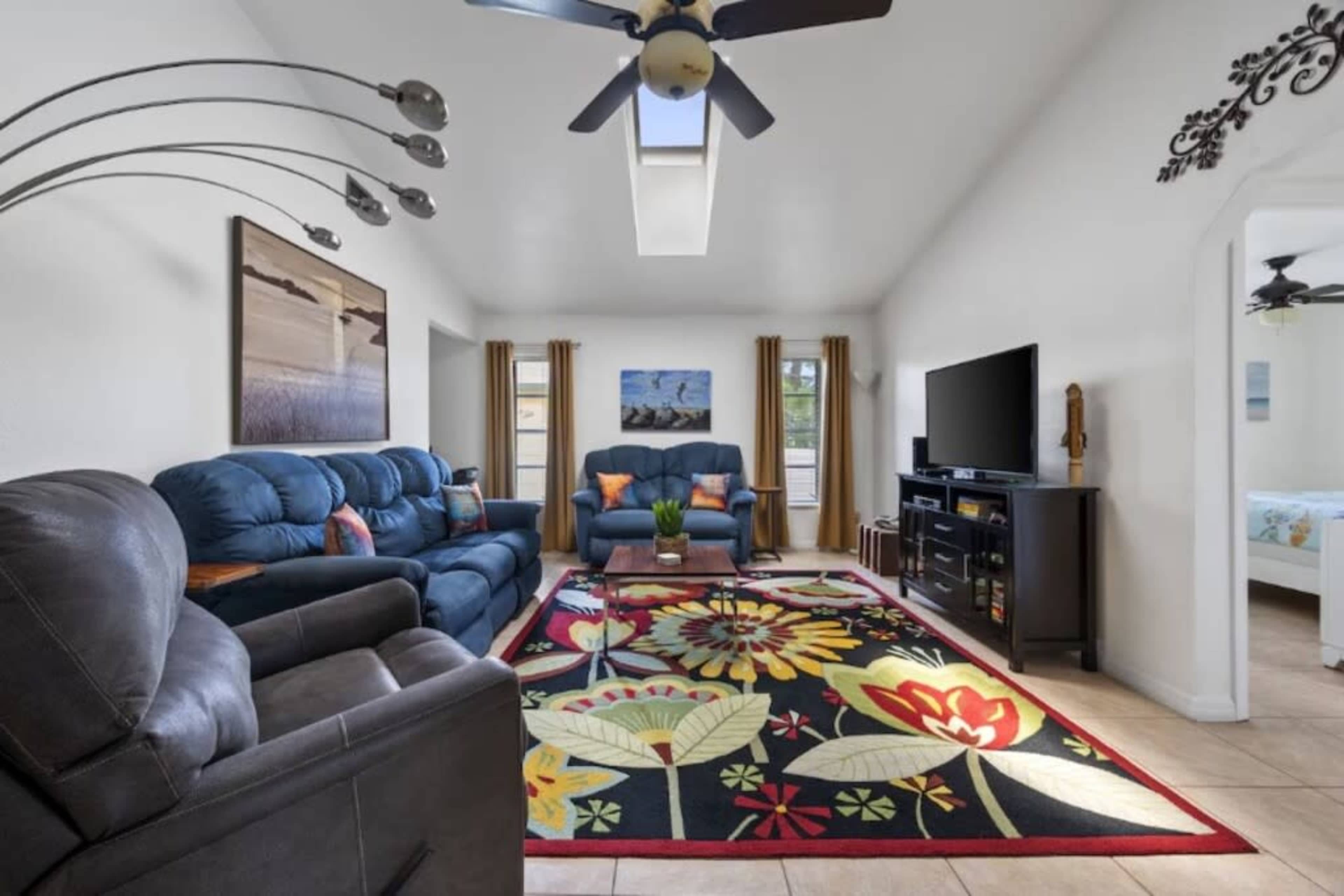 The image shows a living room furnished with blue sofas, a black entertainment unit, and a colorful floral area rug, under a ceiling fan.