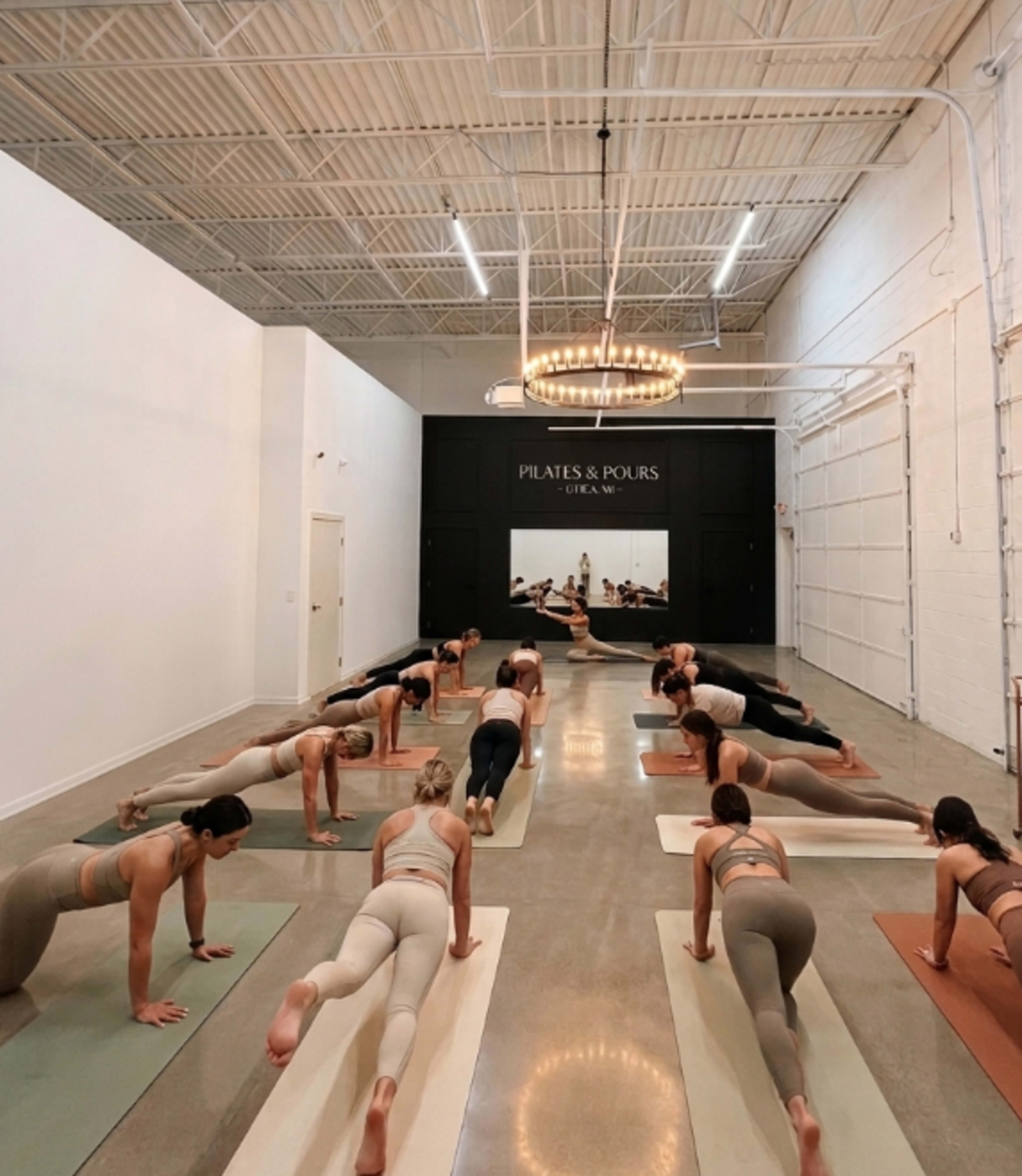 A group of people is practicing Pilates on exercise mats in a well-lit studio with a stylish sign reading "Pilates & Pours" on the wall.
