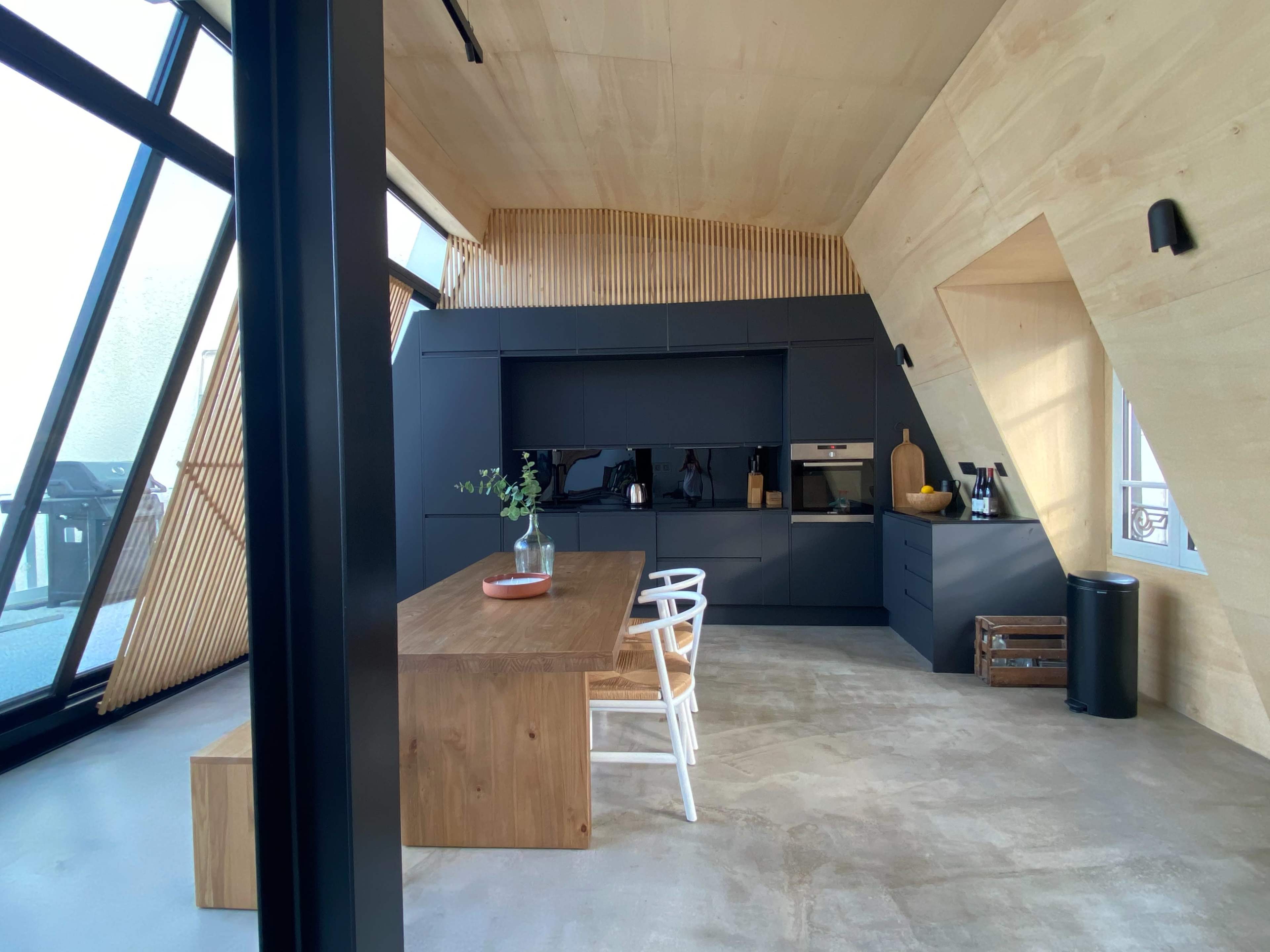 A modern kitchen with a wooden table and black cabinetry, featuring large windows and angled walls.